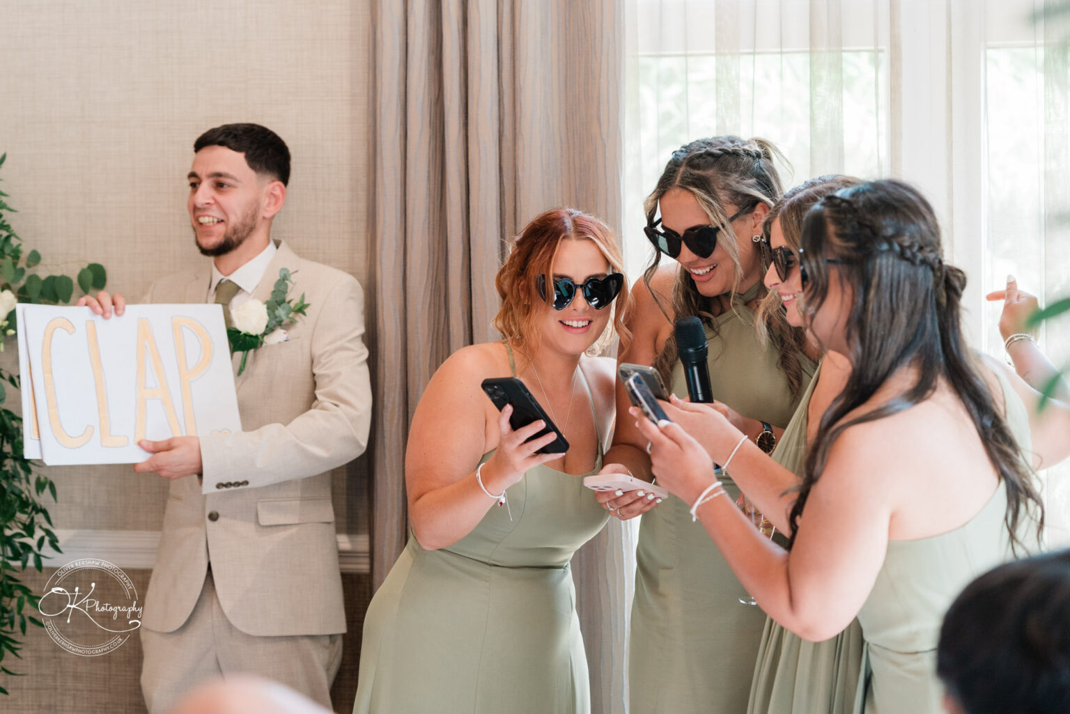 A group of four people, including a man in a light suit holding a sign that says "CLAP," and three women in green dresses, wearing sunglasses and holding phones, appear to be engaged in a fun moment at an event, possibly celebrating a wedding.
