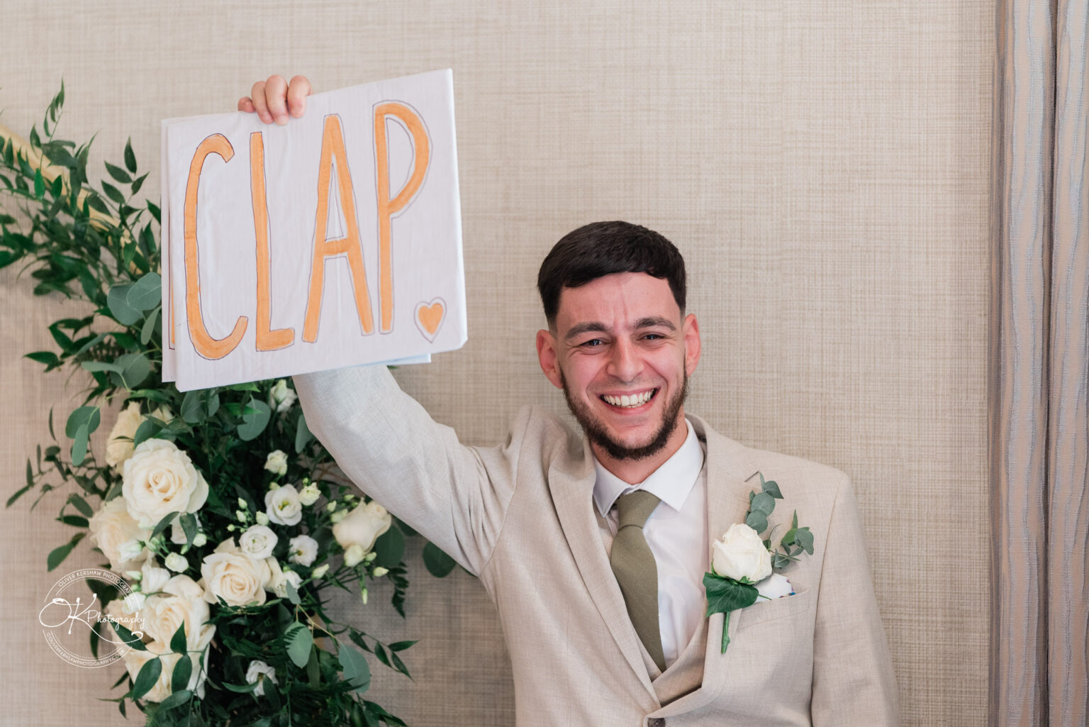 A smiling man in a light-coloured suit holds up a sign that says "CLAP" in orange letters, with a heart underneath. He is standing in front of a backdrop featuring greenery and white flowers.