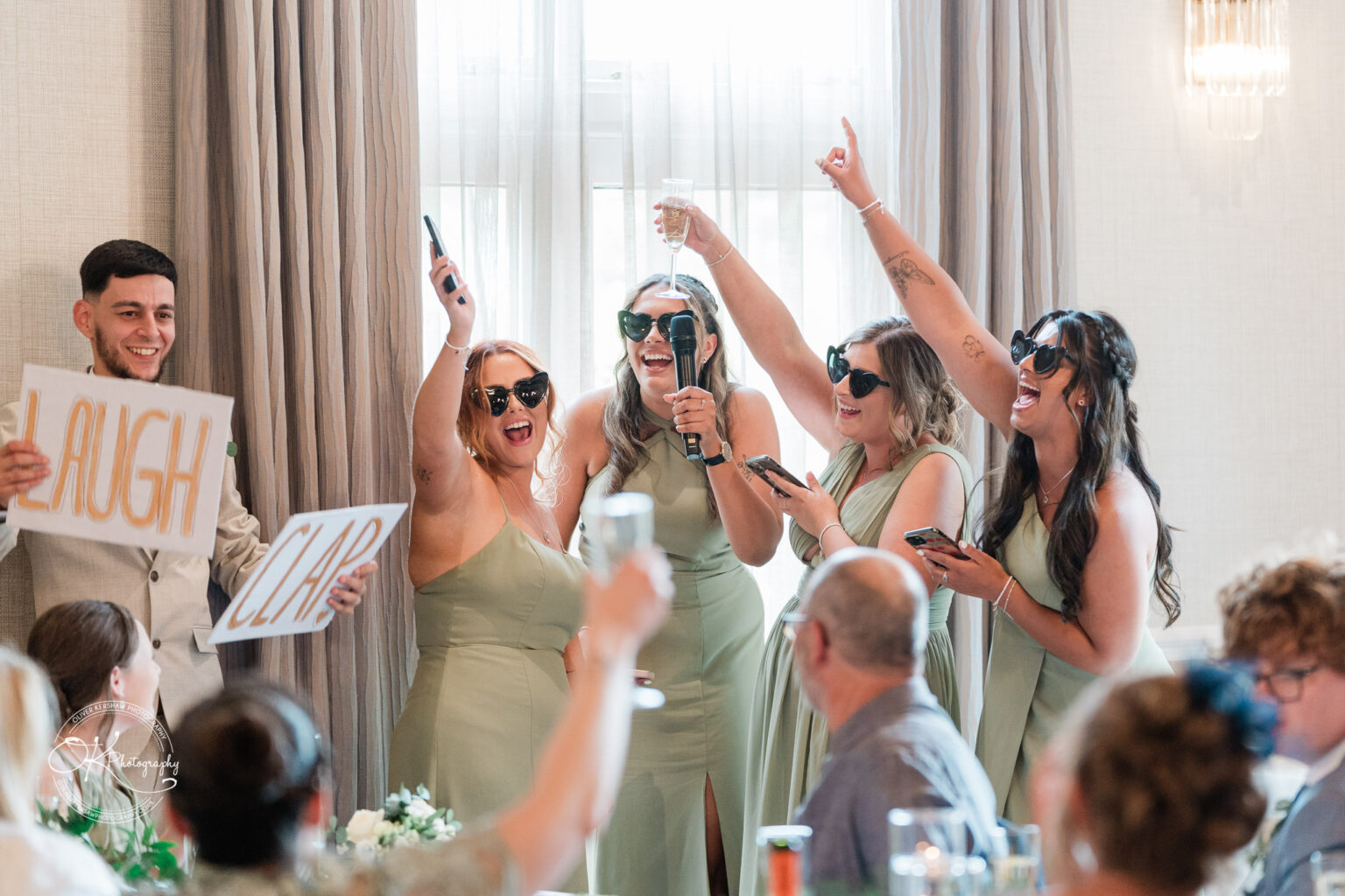 A group of women in green dresses and sunglasses are joyfully celebrating at an event, holding signs that say "LAUGH" and "CLAP," while one woman holds a microphone and another raises a glass. Guests in the foreground are looking towards them.