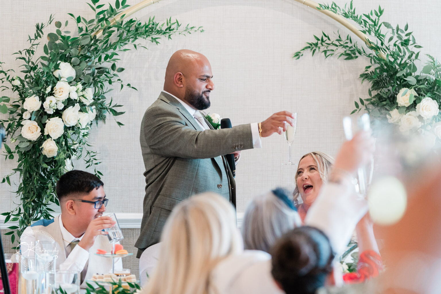 A man in a grey suit raises a glass in a toast during a celebration, while a woman with blonde hair joyfully responds. A boy with glasses is seated nearby, sipping from a glass. The background features floral decorations.