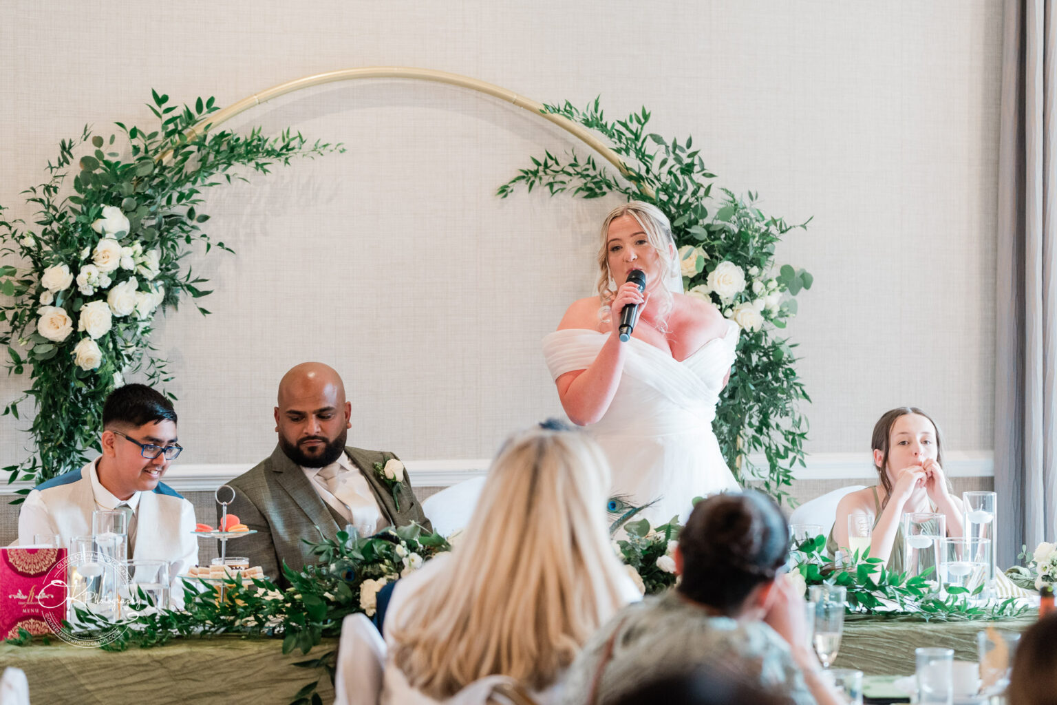 A woman in a white dress speaks into a microphone while seated at a decorated table with greenery and flowers. Beside her are a man and a boy, both looking towards her. In the foreground, a few guests listen attentively, with one woman in a patterned dress visible.