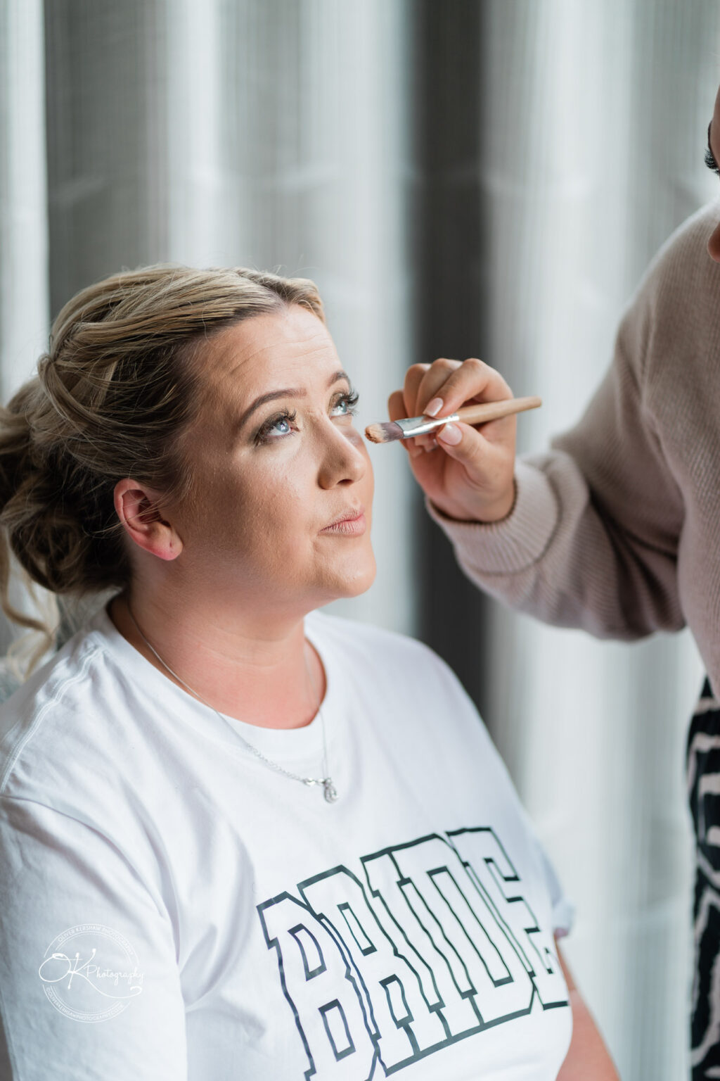 A woman sitting in front of a window is having her makeup applied by another person. She is wearing a white t-shirt with the word "BRIDE" written on it. The makeup artist is using a brush to apply makeup near the woman's eyes.