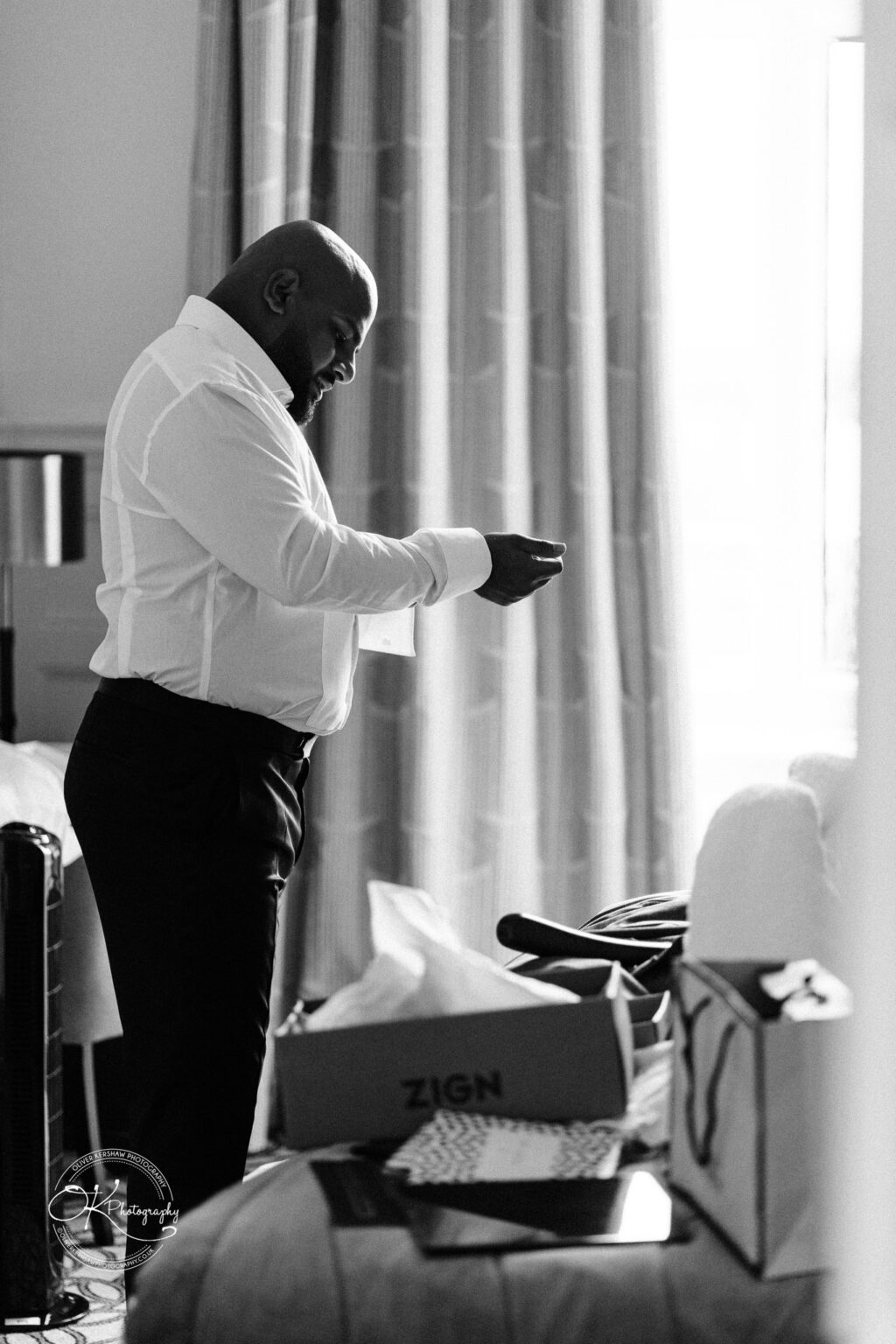 A man in a white shirt and black trousers stands in profile, looking down at a small object in his hands, surrounded by scattered items on a bed and soft light coming through a nearby window.