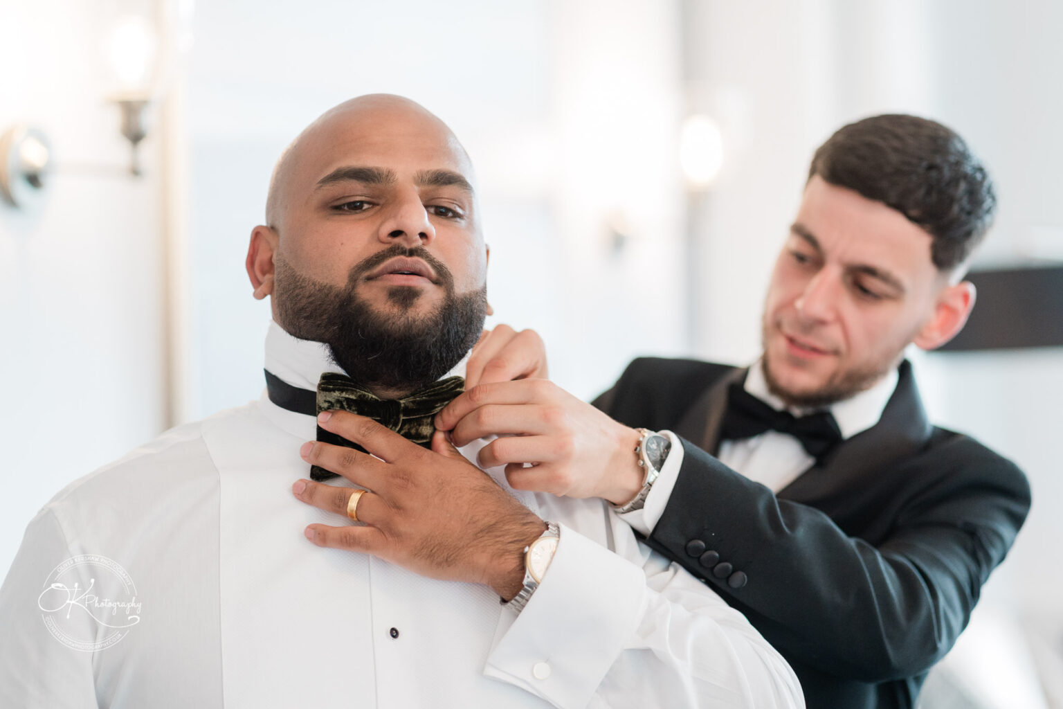 A man in a white shirt is having a bow tie adjusted by another man in a black tuxedo. The setting appears to be a bright and elegantly decorated room.