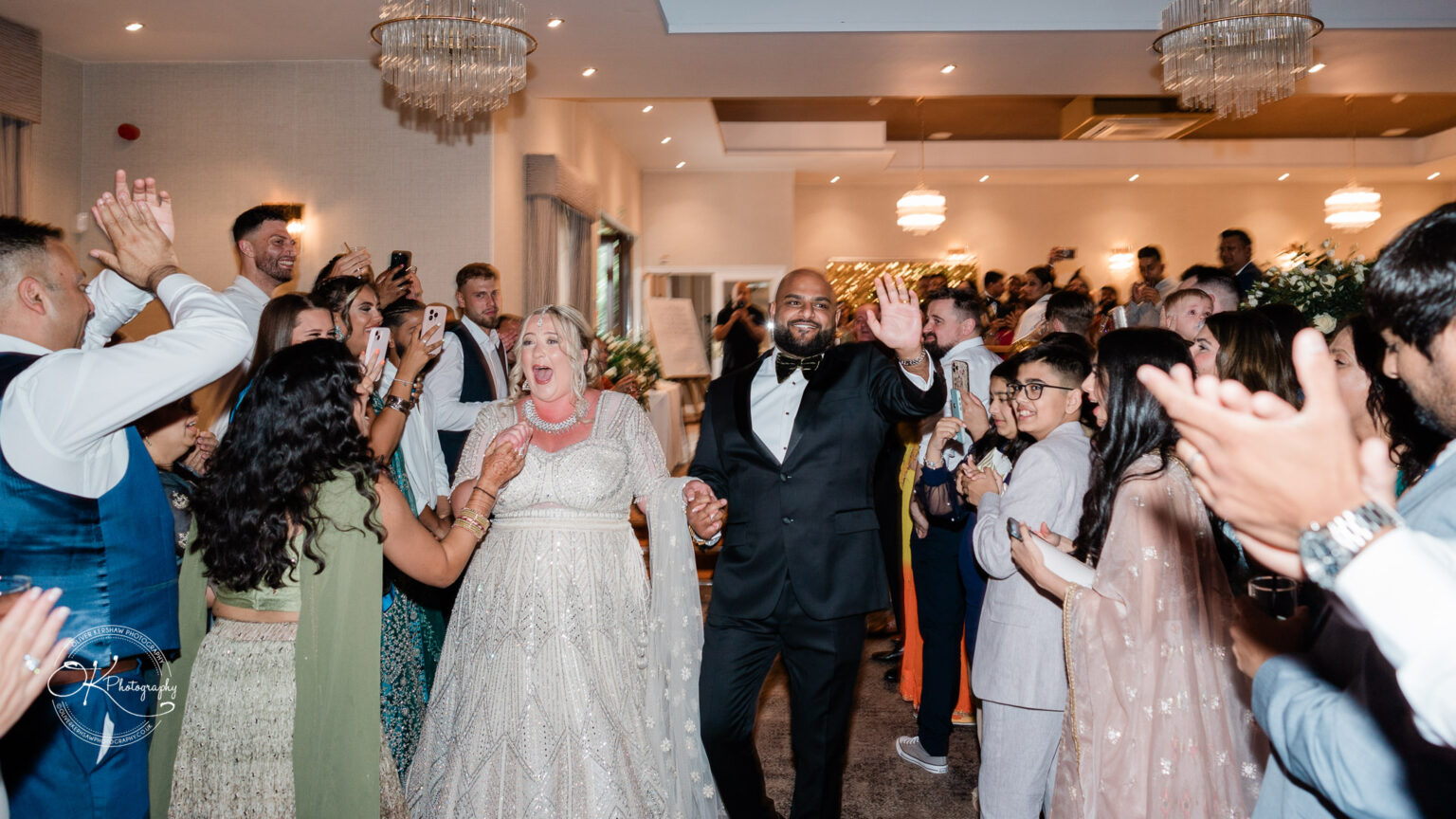 A bride and groom joyfully enter a celebration surrounded by smiling guests clapping and taking photos, while chandeliers illuminate the festive atmosphere.