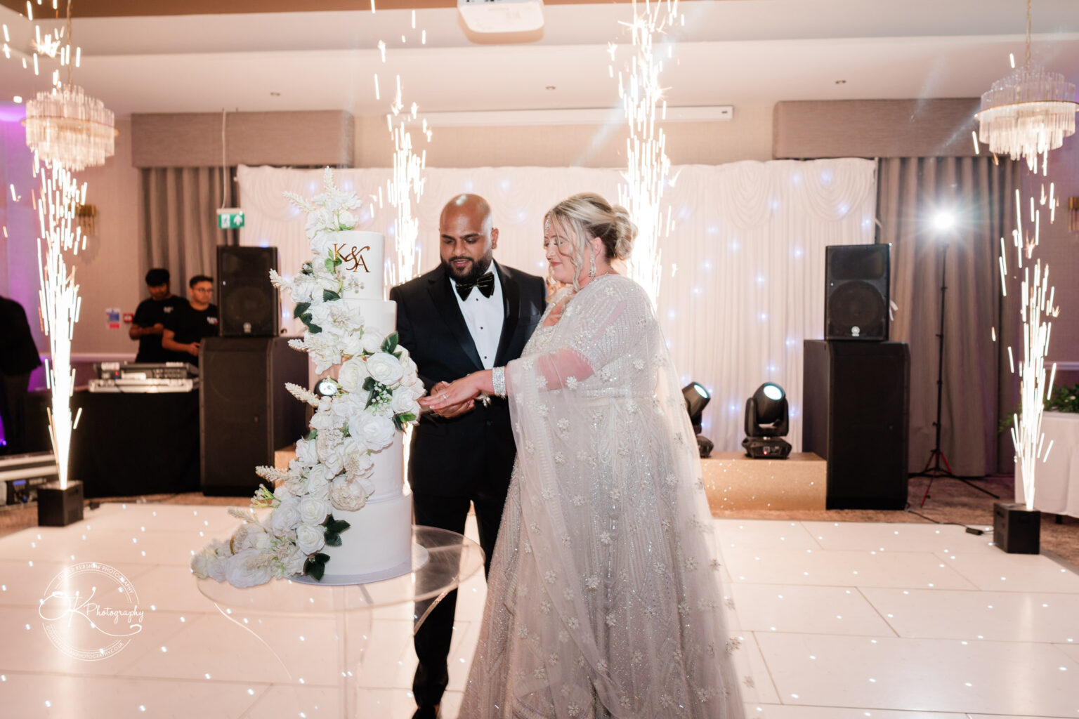 A man in a tuxedo and a woman in a sparkling, beaded gown stand together by a three-tier wedding cake adorned with flowers, while sparklers erupt around them in a festive setting.