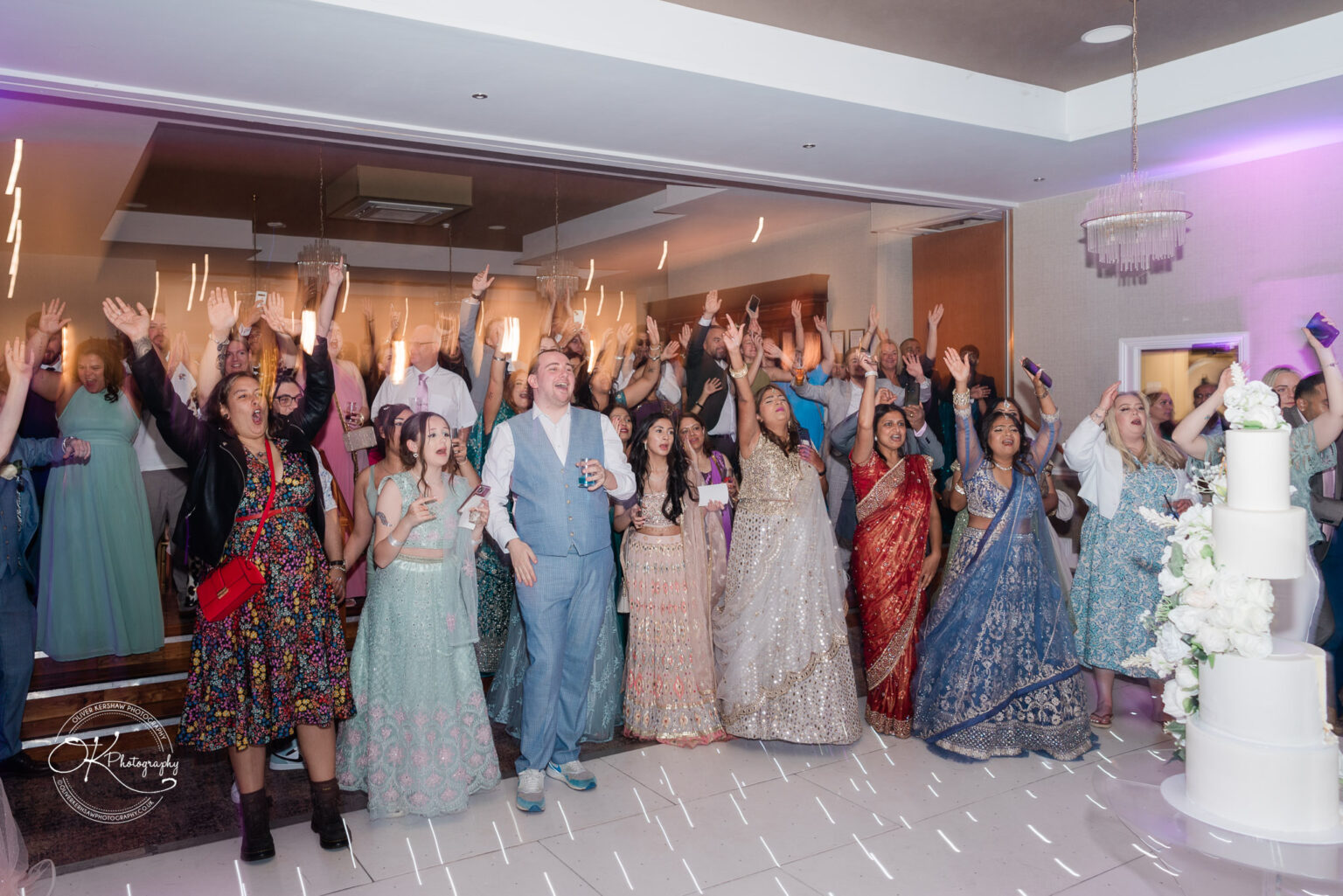 A lively group of guests at a celebration, with many people raising their hands in excitement, dressed in a variety of formal attire, including ethnic outfits. In the foreground, a multi-tiered wedding cake is decorated with white flowers.