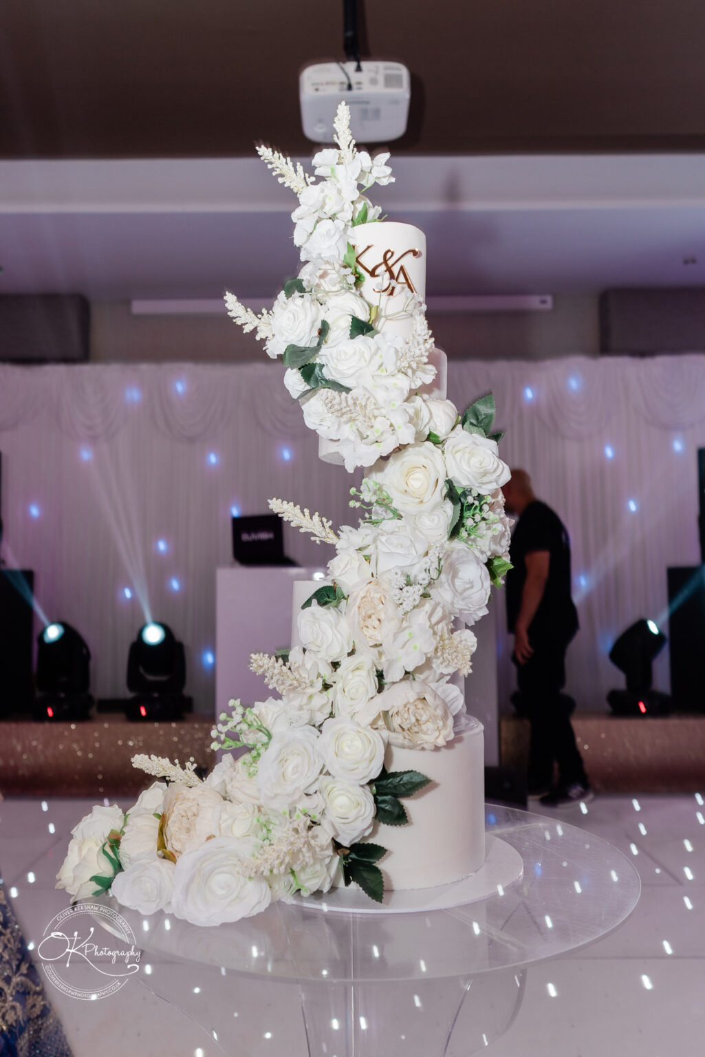 A tall, white wedding cake adorned with cascading white flowers and greenery, featuring the letters "S&A" in gold on the top tier, set against a softly lit event backdrop.