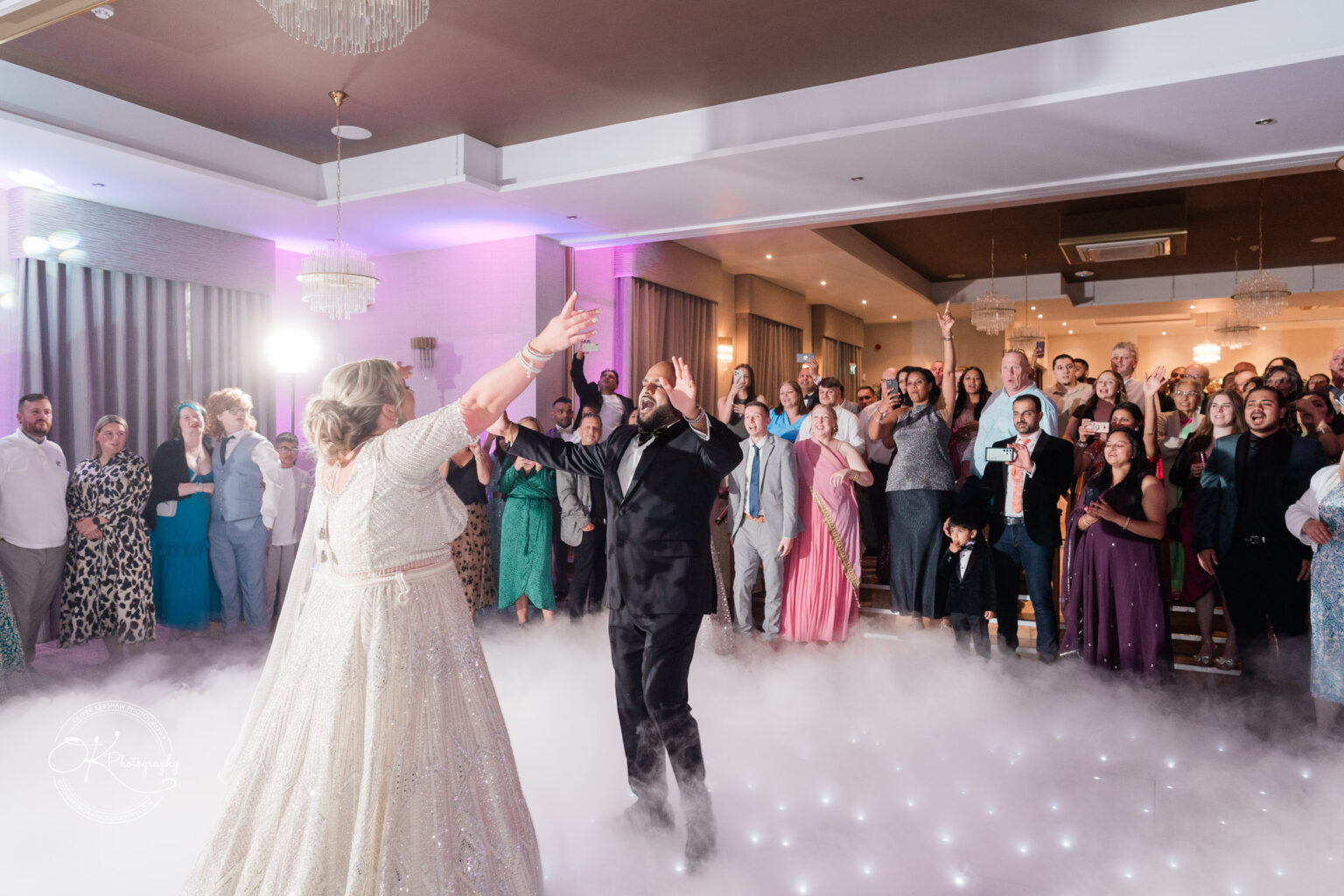 A couple is dancing on a cloud-like floor, surrounded by an enthusiastic crowd in formal attire, celebrating at a wedding reception. The room is elegantly decorated with soft lighting and chandeliers.