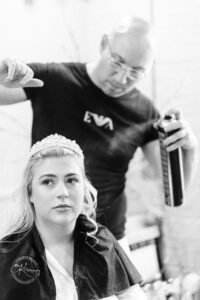 A woman with long blonde hair wears a tiara and a black cape while sitting in a salon chair, looking thoughtfully ahead. A stylist stands behind her, spraying hair products from a canister. The image is in black and white.