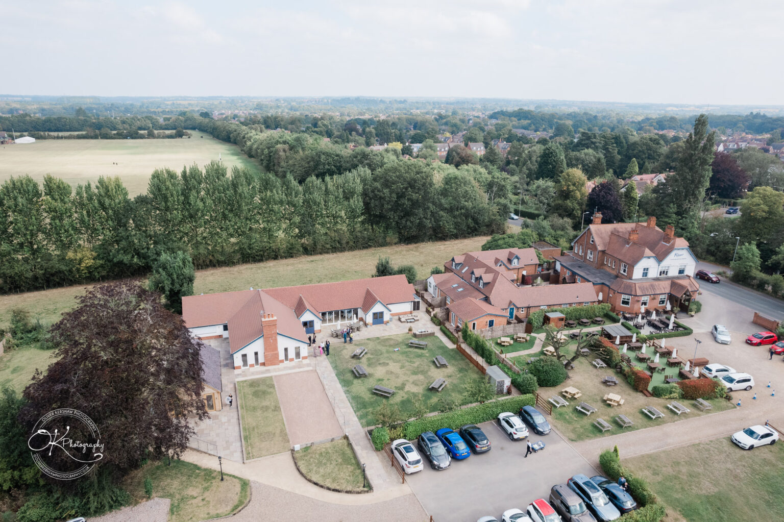 Aerial view of a small village area featuring several buildings with red roofs, a lawn, and parking spaces. In the foreground, a landscaped area with benches and pathways is visible, alongside a larger grassy field and treed areas in the background.