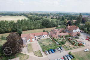 Aerial view of a small village area featuring several buildings with red roofs, a lawn, and parking spaces. In the foreground, a landscaped area with benches and pathways is visible, alongside a larger grassy field and treed areas in the background.