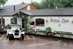 A vintage white car is parked in front of a quaint building with the sign "The Royal Oak, Kirby Muxloe." The exterior is adorned with greenery and floral decorations, while the ground is wet, indicating recent rain.