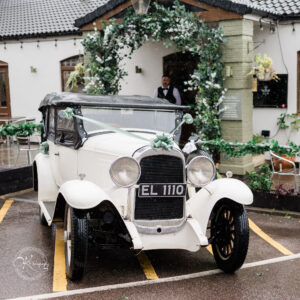 A vintage white car with a black convertible top, decorated with green ribbons, parked in front of a building adorned with greenery and flowers. A person in formal attire is visible in the background.