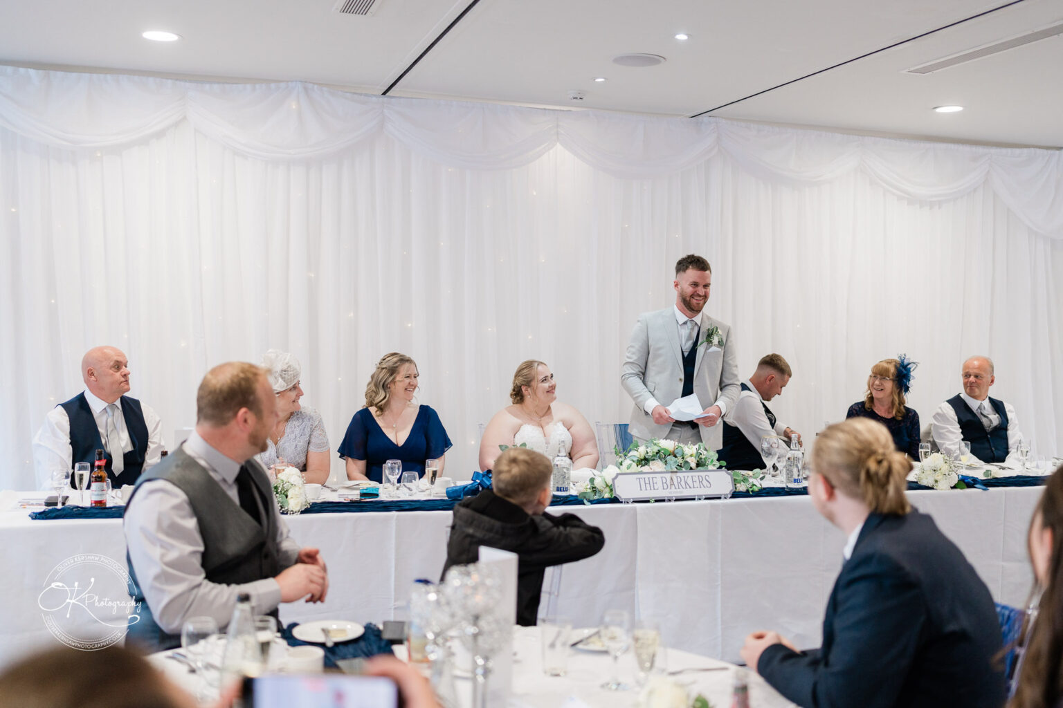 A wedding reception with a man in a light suit speaking at a head table, surrounded by guests and decorations.