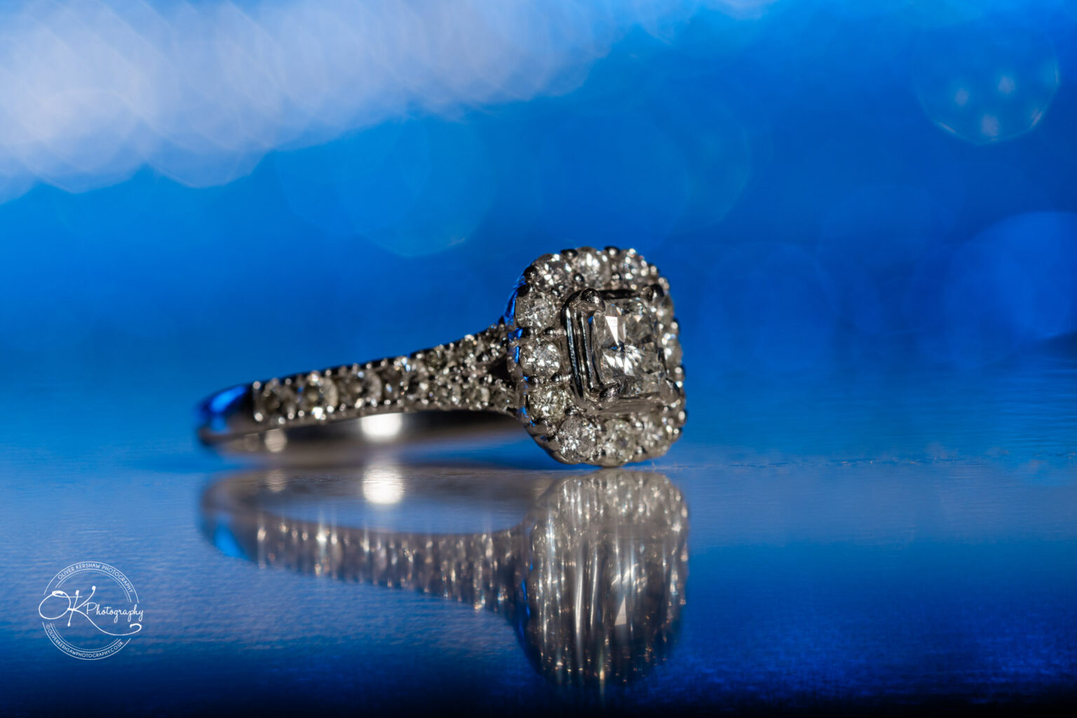 A sparkling diamond ring with a square-shaped centre stone, surrounded by smaller diamonds, sitting on a reflective surface against a blue bokeh background.