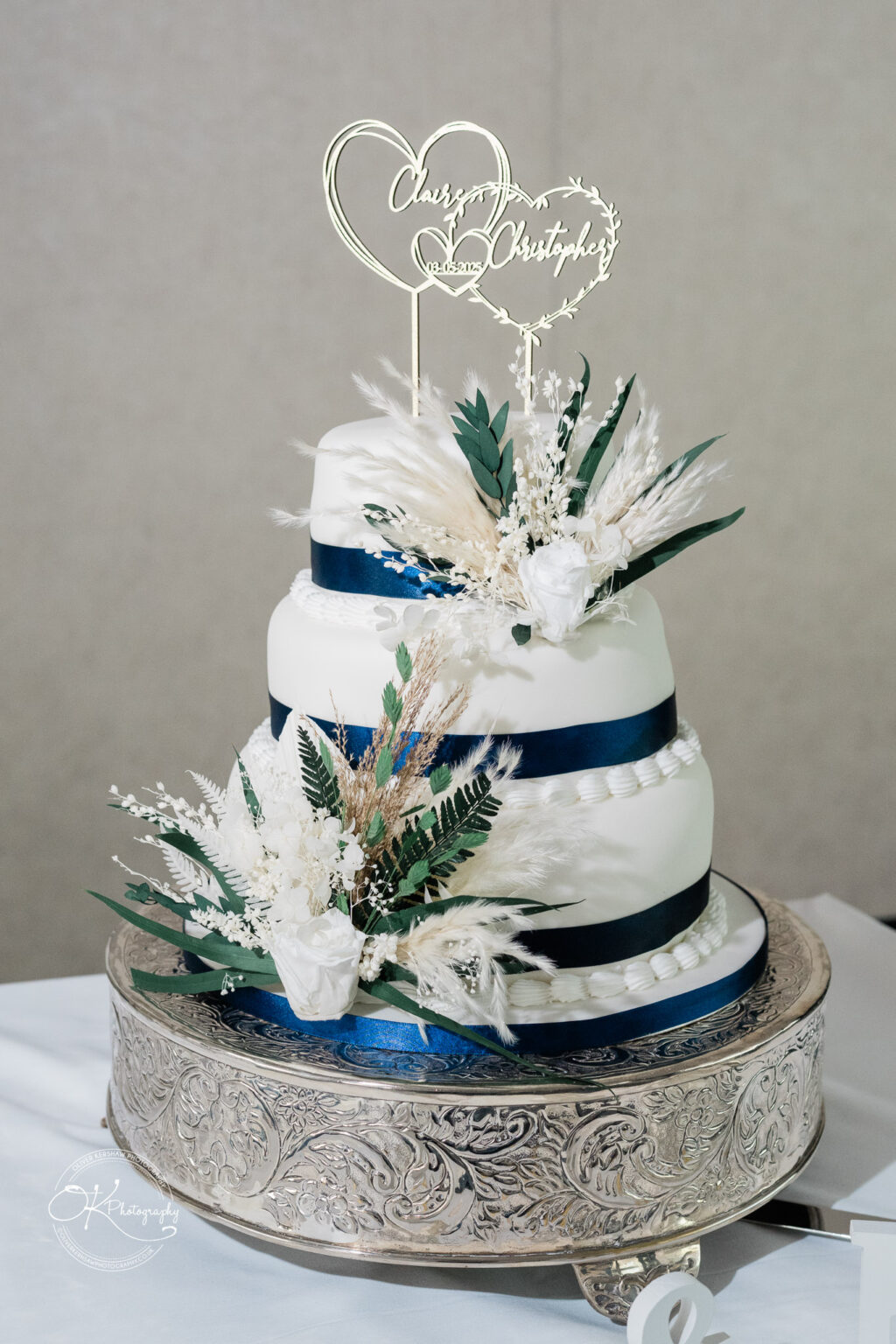 A three-tier wedding cake adorned with navy ribbon, floral decorations, and a heart-shaped topper with the names "Chase" and "Christopher."