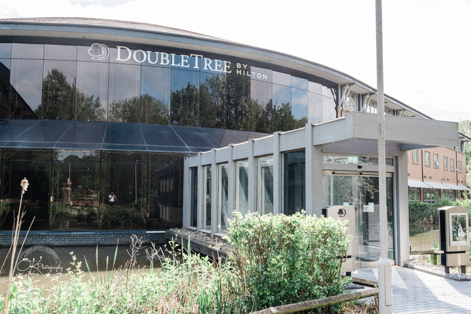 Modern hotel building with large glass windows and the "DoubleTree by Hilton" sign, surrounded by greenery and a canal.