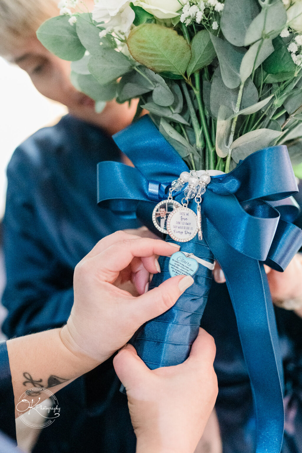 A person adjusts a blue satin ribbon around a bouquet, which has charms and a small note attached, while another figure observes in the background.