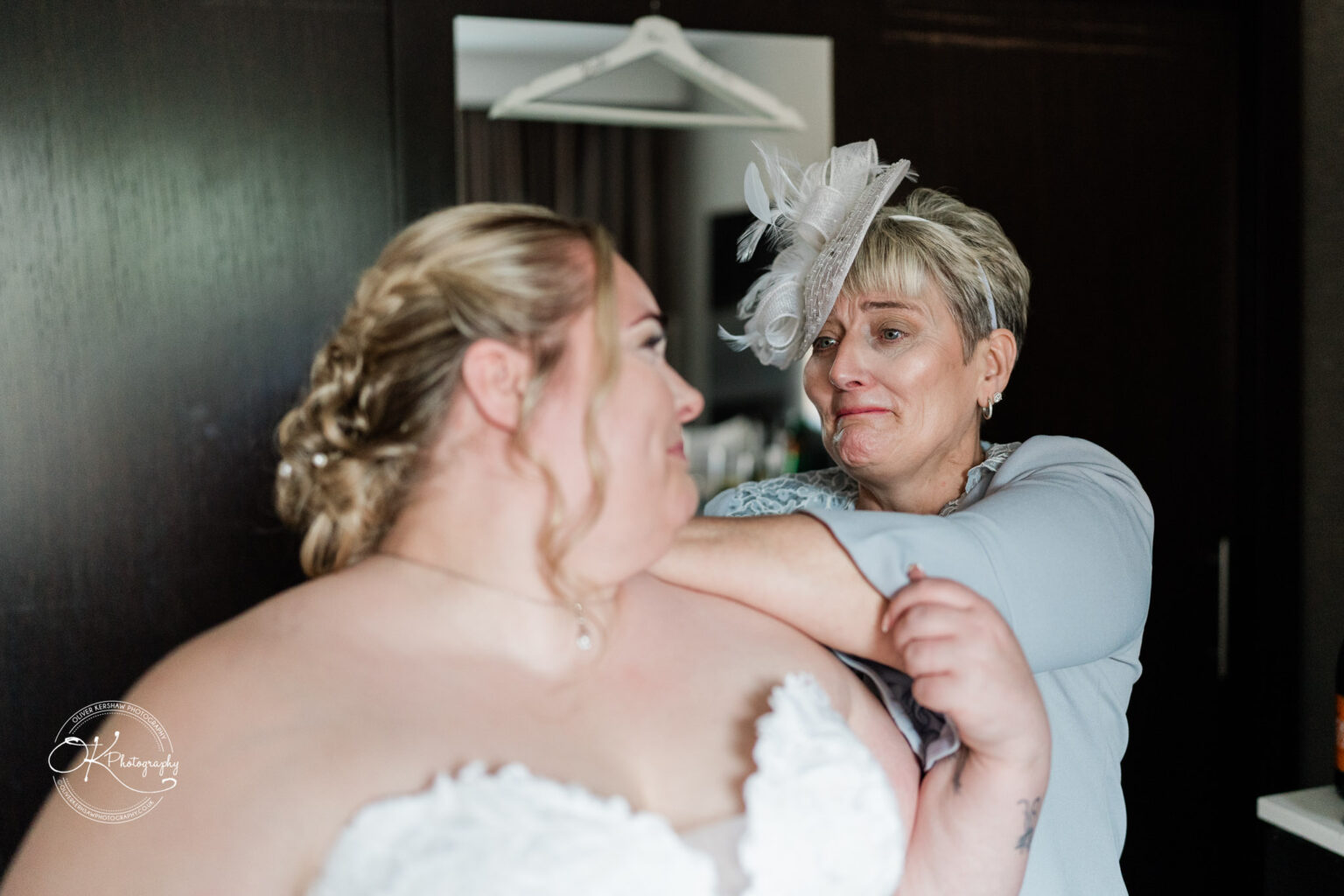 A bride in a strapless wedding dress smiles at her mother, who is wearing a fascinator, as they share an emotional moment in a room.