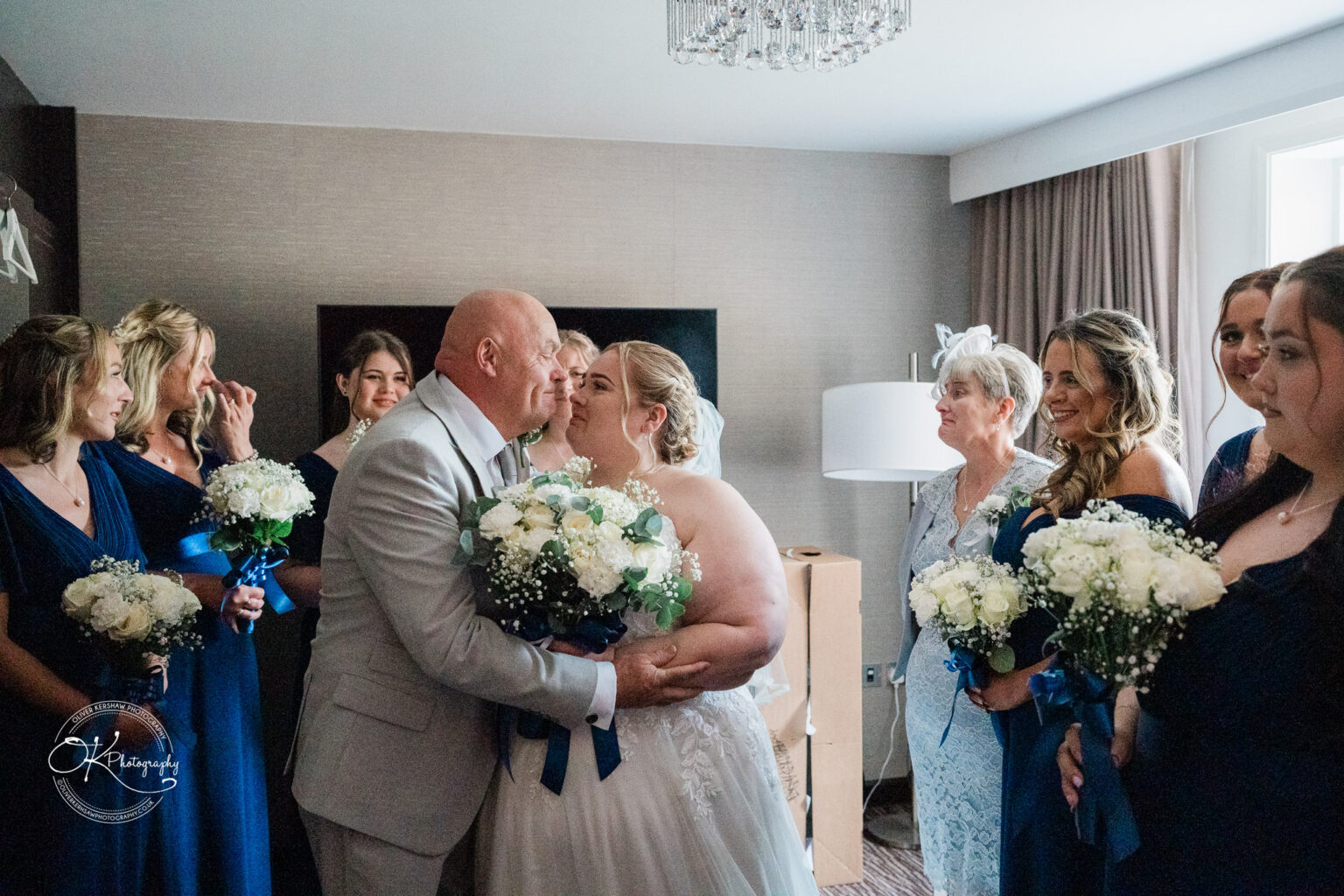 A bride and groom share a kiss in a hotel room surrounded by bridesmaids in blue dresses and a woman in a light-coloured outfit.