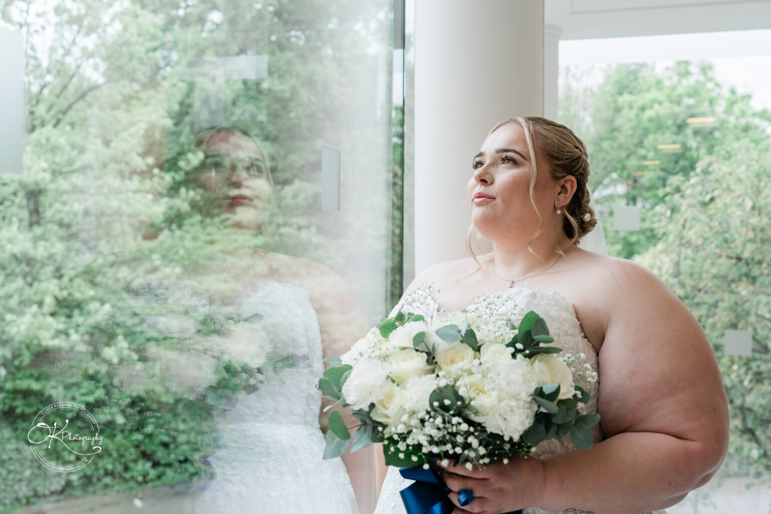 A bride in a white wedding dress stands by a large window, holding a bouquet of white flowers with greenery, gazing thoughtfully outside.
