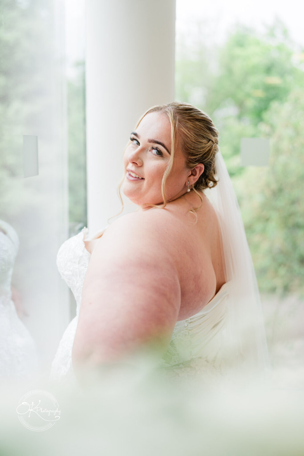 A bride with long hair and a veil smiles while looking over her shoulder, standing by a large window with greenery in the background.