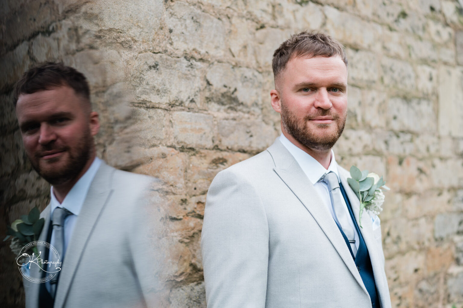 A man in a light grey suit and tie stands beside a stone wall, with a soft reflection of him visible on the surface.