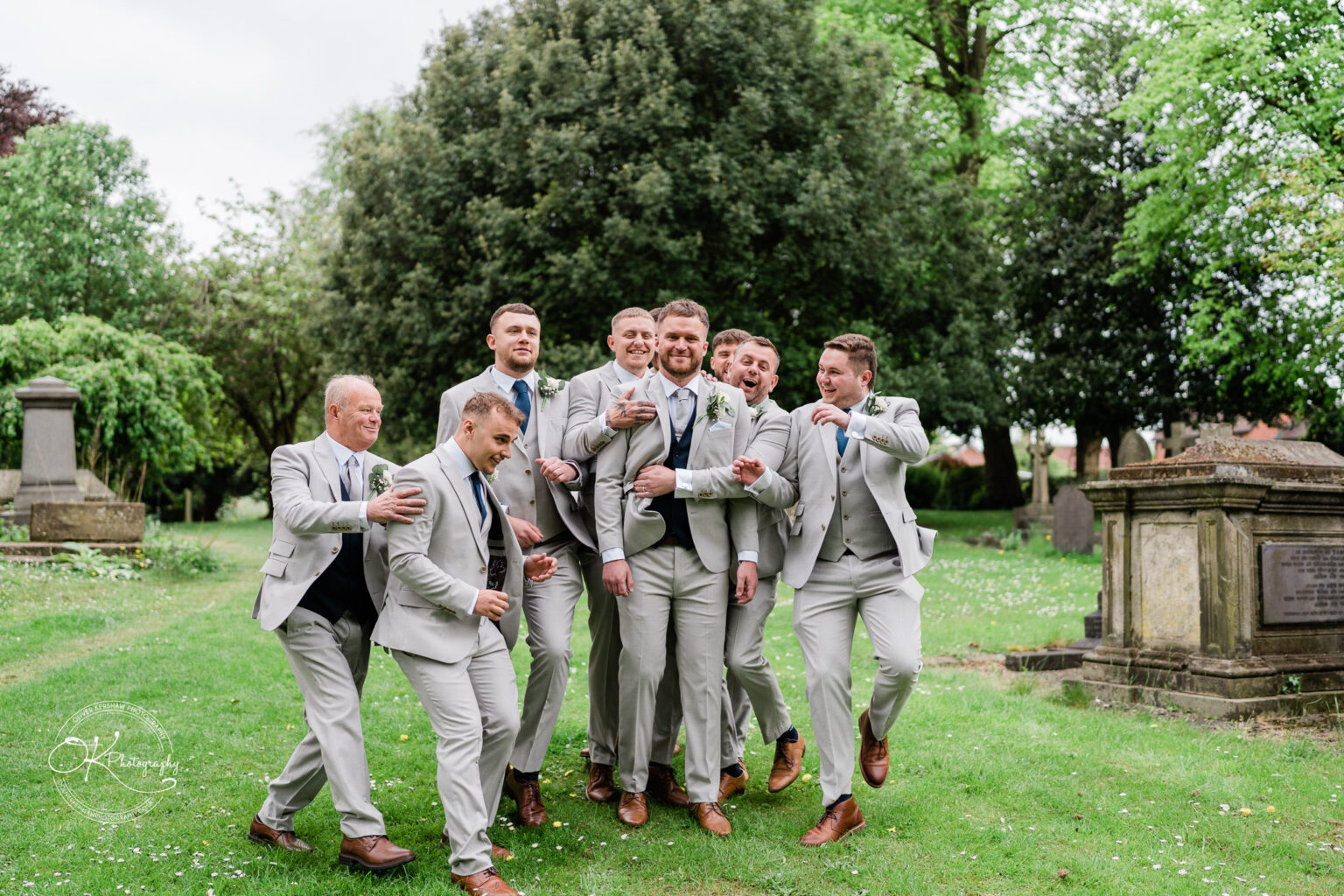 A group of eight men in light grey suits and blue ties joyfully embrace and pose playfully in a grassy cemetery setting.