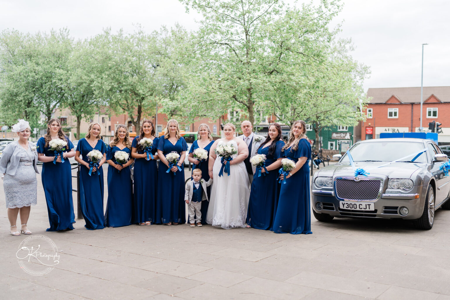 A group of bridesmaids in blue dresses stands with a bride in white, holding bouquets, beside a luxury car.