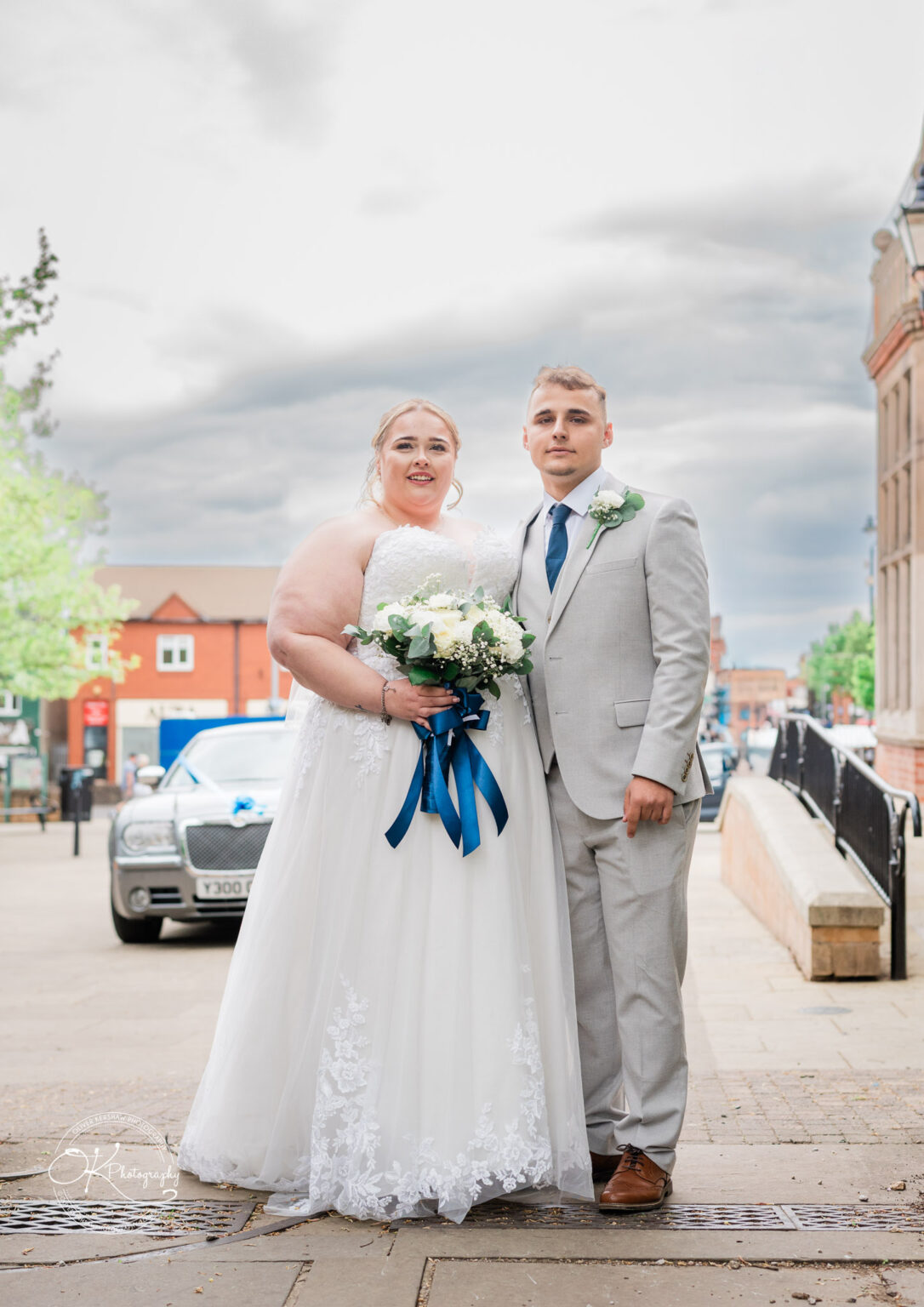 A newlywed couple stands together outdoors, the bride in a white wedding dress holding a bouquet with blue ribbons, and the groom in a light grey suit.