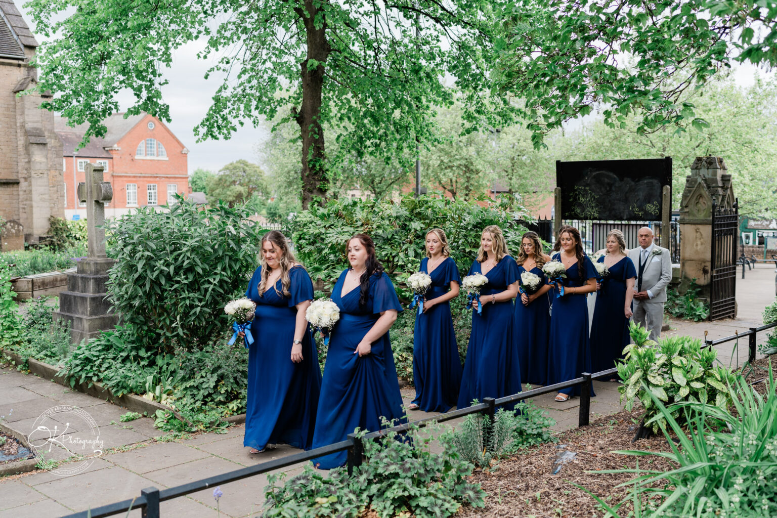 A group of bridesmaids in long navy dresses walks outdoors, holding bouquets, surrounded by greenery and a scenic backdrop.
