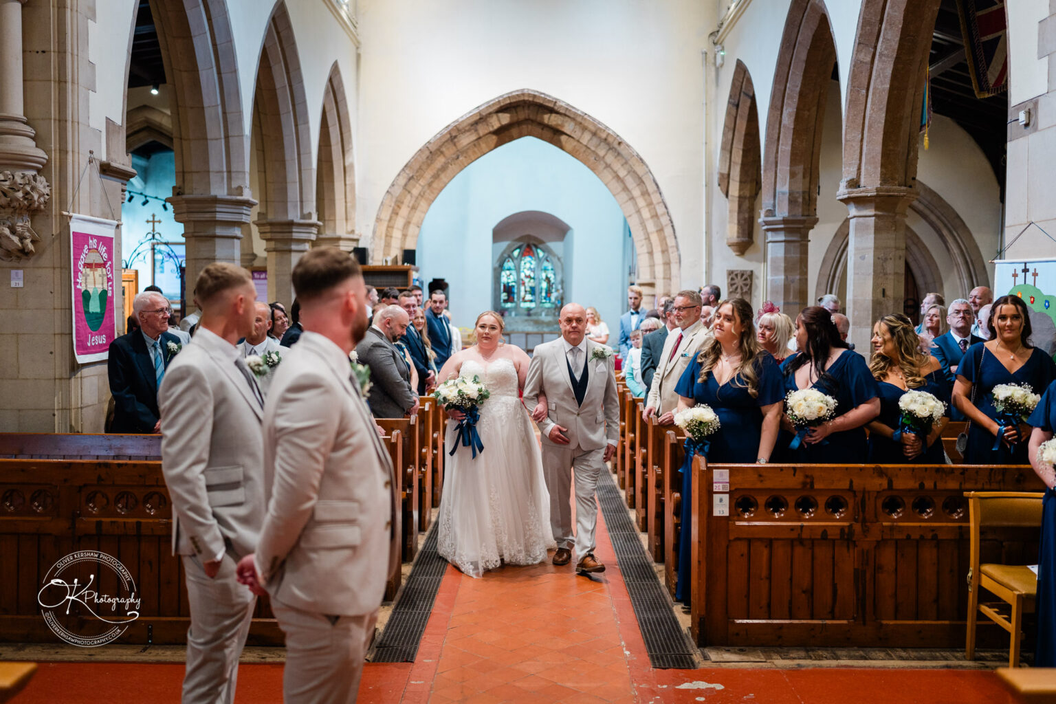 A bride in a white dress walks down the aisle with her father, surrounded by wedding guests seated in a church.