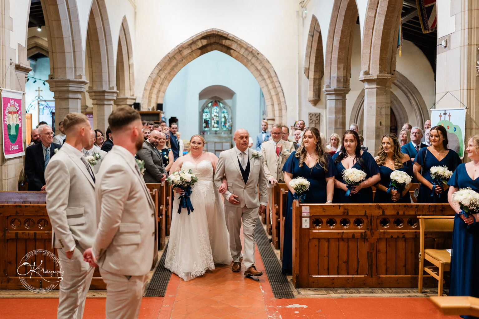 A bride in a white dress walks down the aisle accompanied by a man, while guests look on in an elegant church setting.