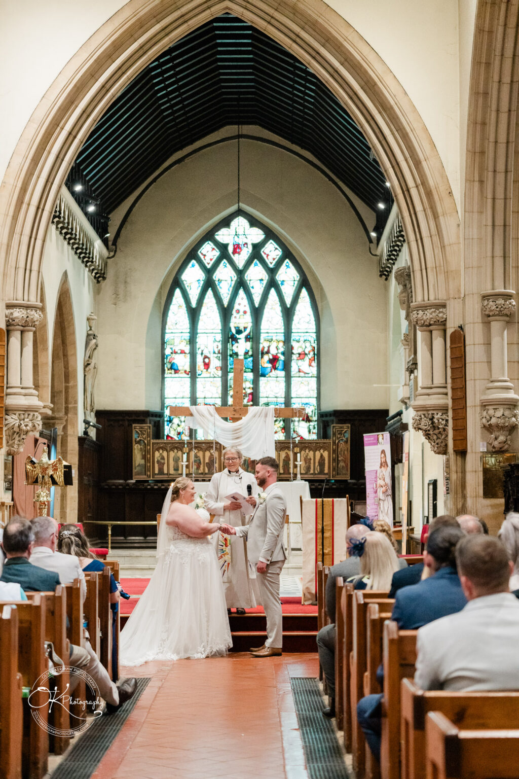 A bride and groom exchange vows in a church, with an ornate stained glass window and guests seated in pews.
