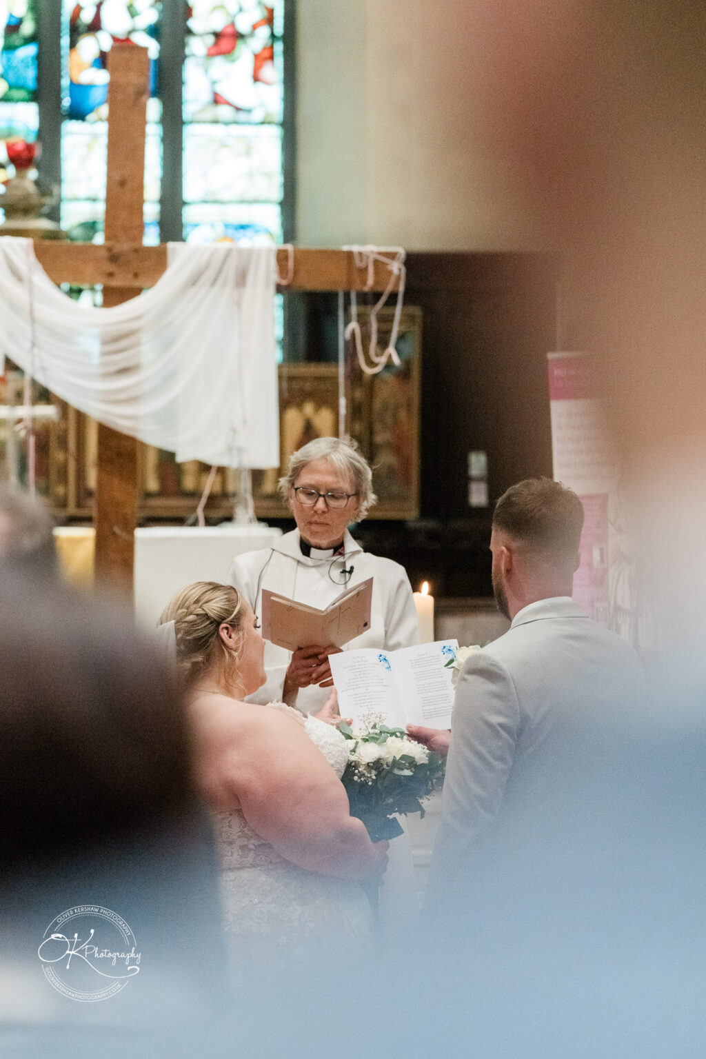 A bride in a wedding dress stands holding a bouquet while reading vows to a groom, with an officiant in the background.
