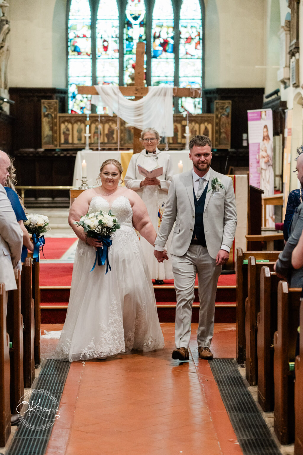A bride in a white gown and groom in a light grey suit hold hands, walking down the aisle of a church, with stained glass windows in the background.