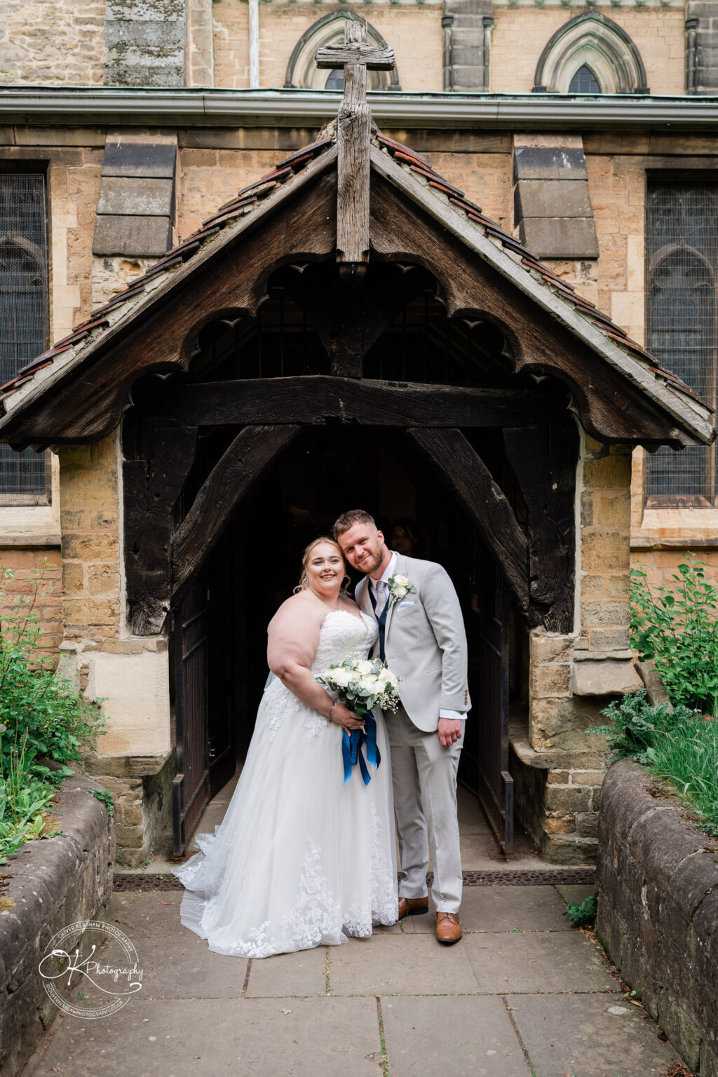 A bride in a white gown and a groom in a light grey suit pose happily together at the entrance of a rustic stone church.