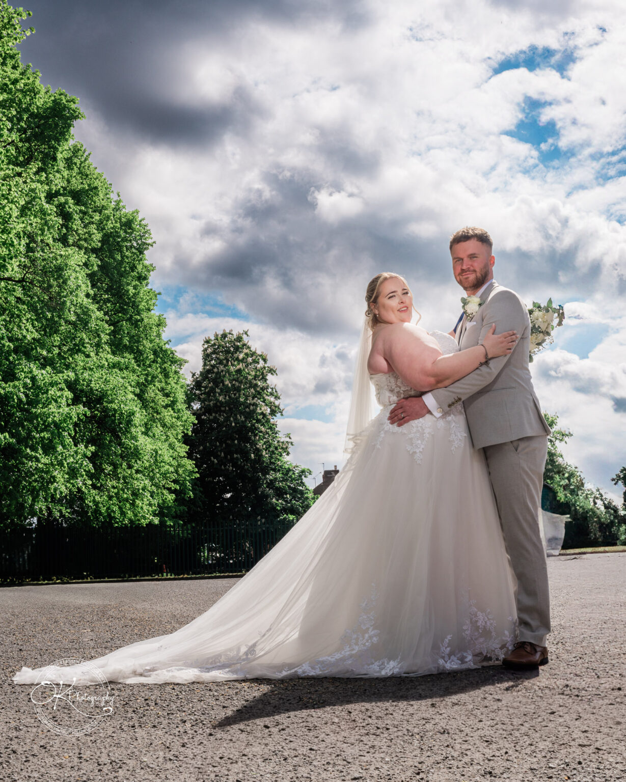 A bride in a flowing white gown embraces a groom in a light grey suit, with a backdrop of fluffy clouds and green trees.