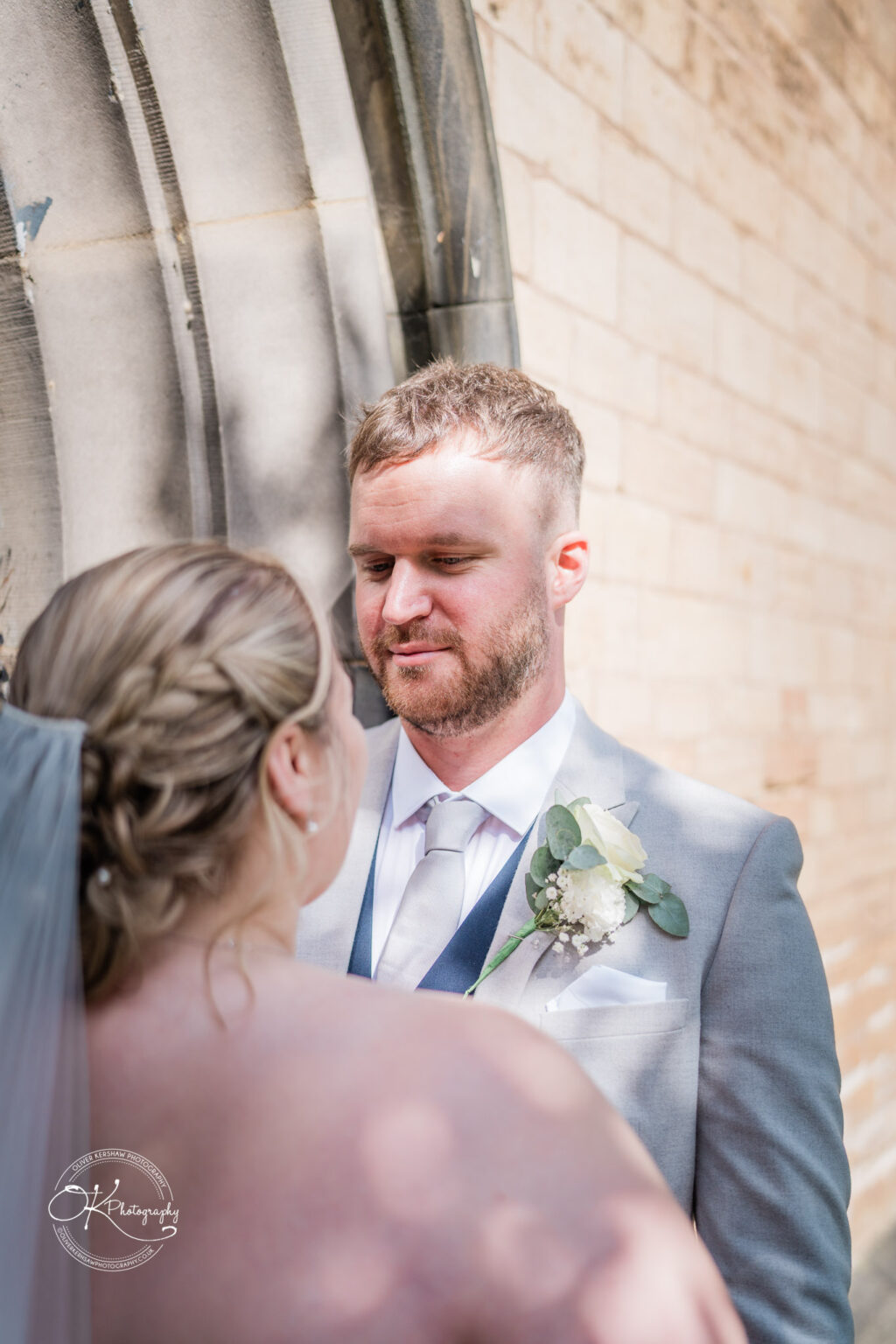 A groom in a grey suit gazes affectionately at his bride, who is partially visible with a veil, against a stone wall backdrop.