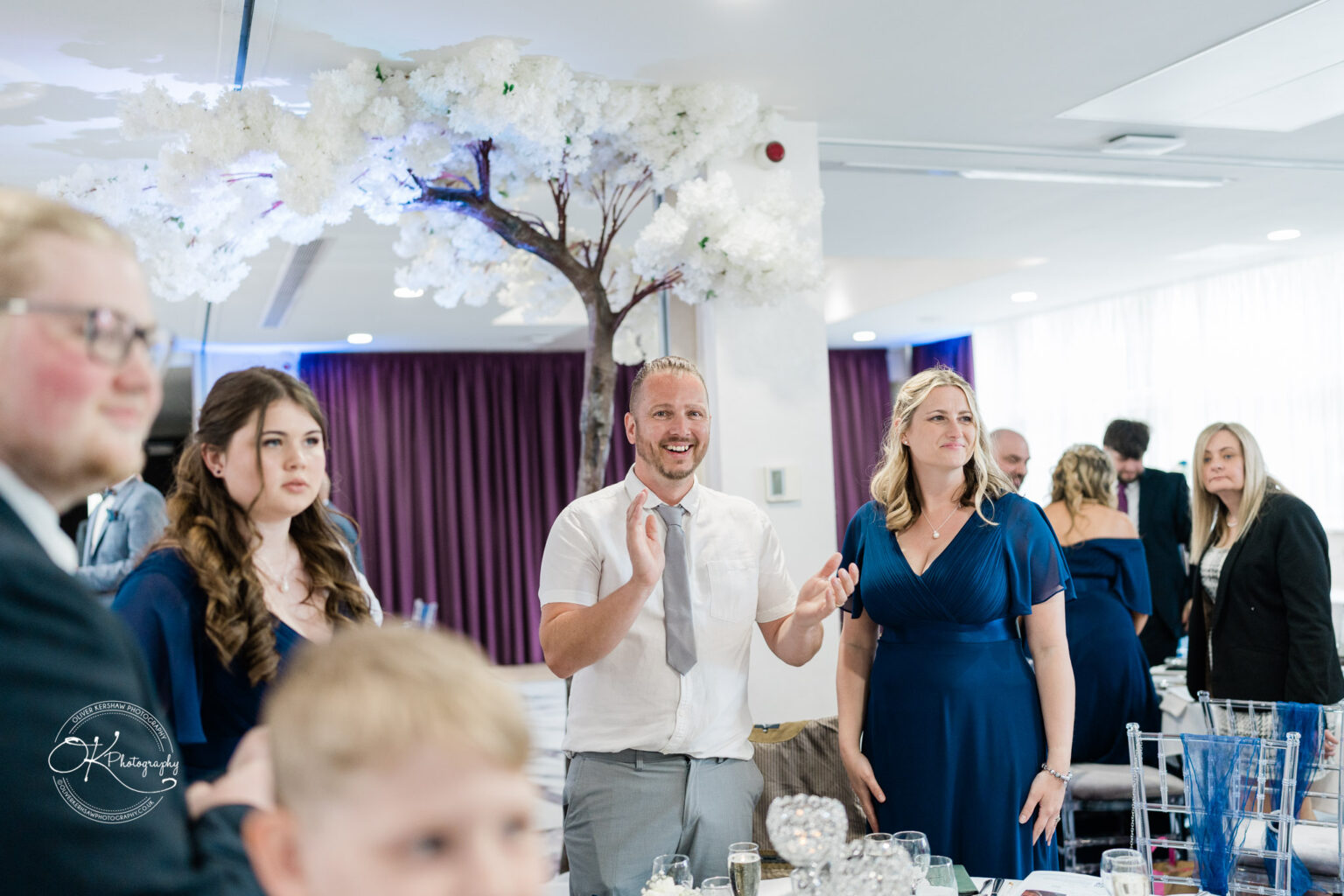 A group of people at a celebration, with a man in a white shirt clapping and smiling, while women in blue dresses look on. A decorative tree with white flowers is visible in the background.