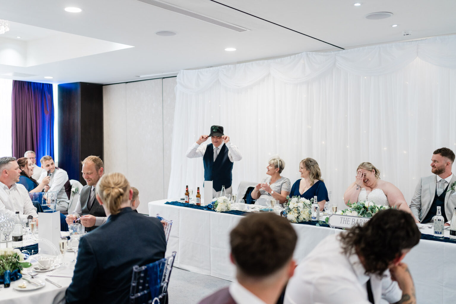 A group of guests seated at a wedding reception, with a man in a vest speaking humorously while wearing a cap, and women in formal attire listening.