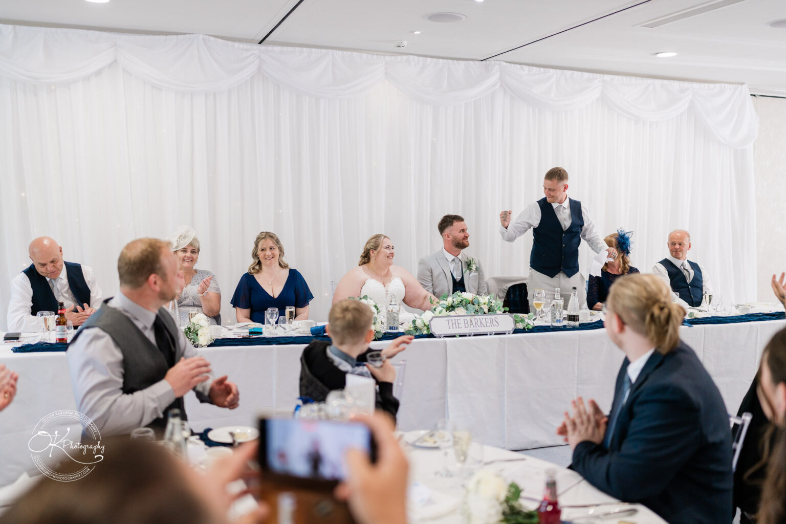 A group of people seated at a long table, celebrating with clapping, while one person stands up to deliver a speech.