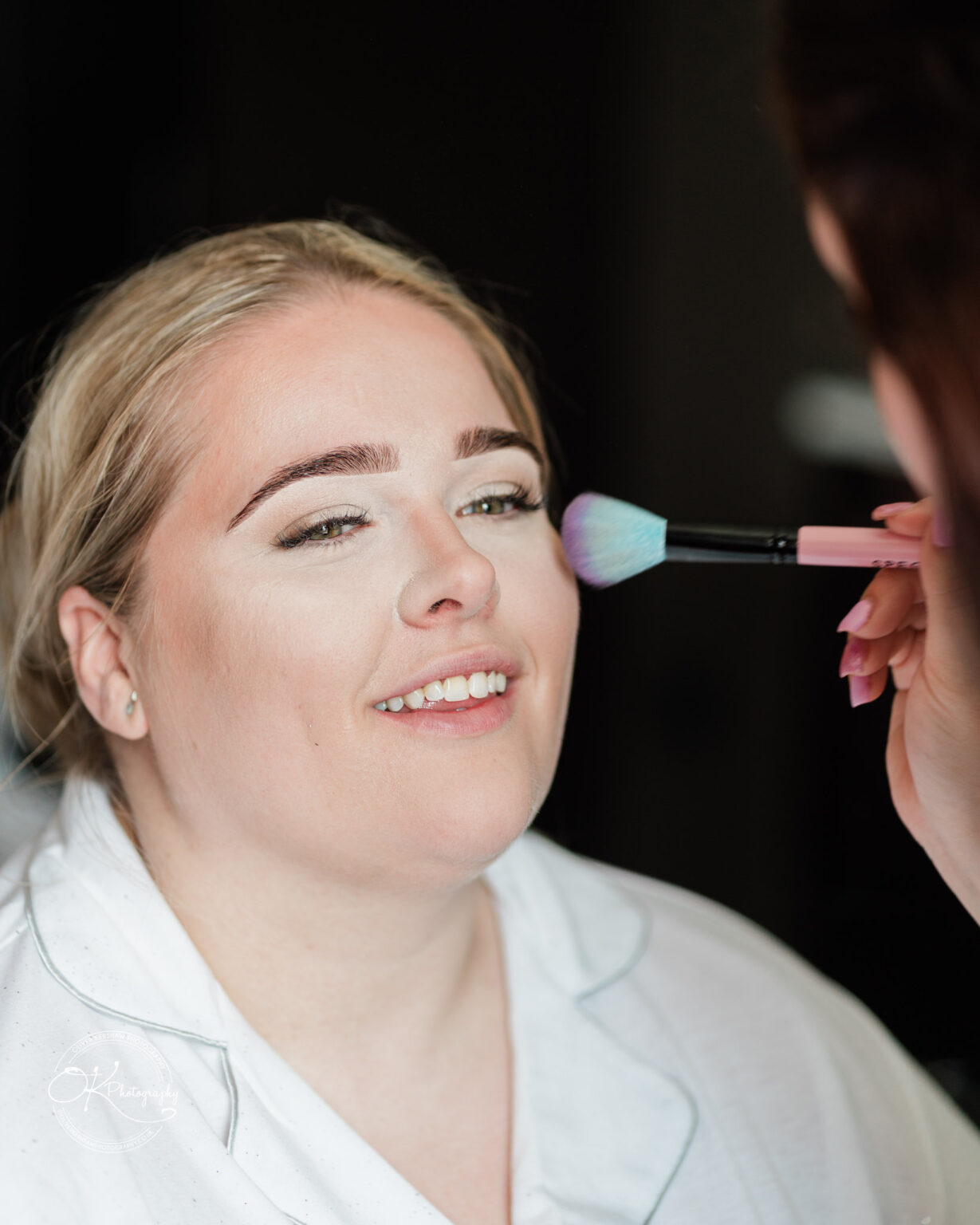 A woman in a white robe smiles as a makeup artist applies makeup to her face with a brush.