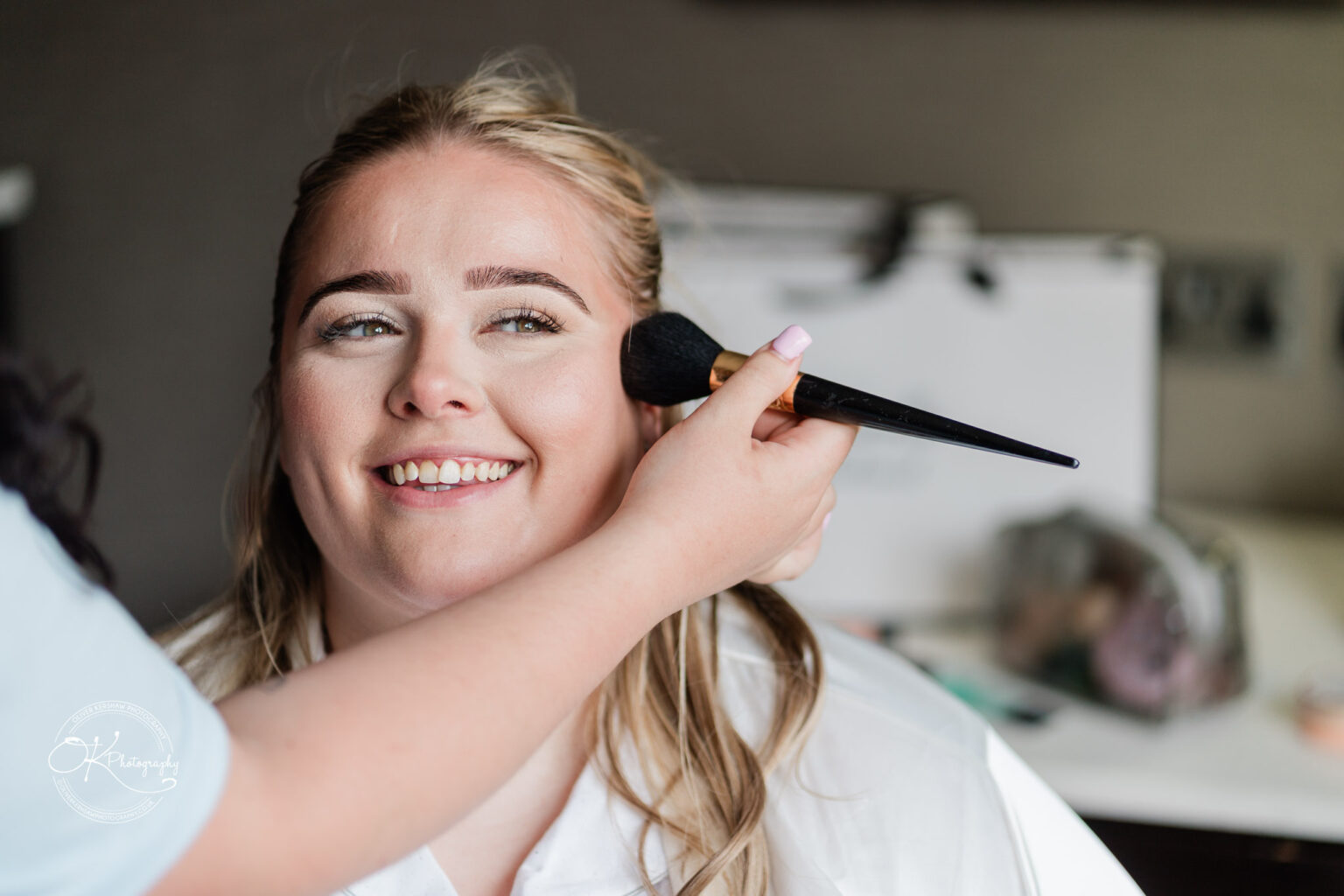A smiling woman sitting in a chair is having her makeup applied by another person, with a soft-focus background featuring cosmetics.