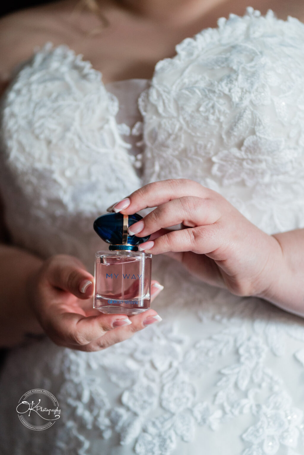 A person holds a small perfume bottle with a dark blue cap, dressed in an intricately detailed white wedding gown.
