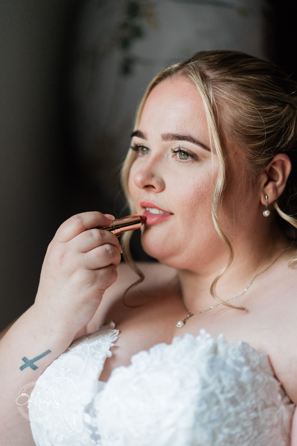 A bride applying lipstick in a close-up shot, wearing a strapless white wedding dress with soft waves in her hair.