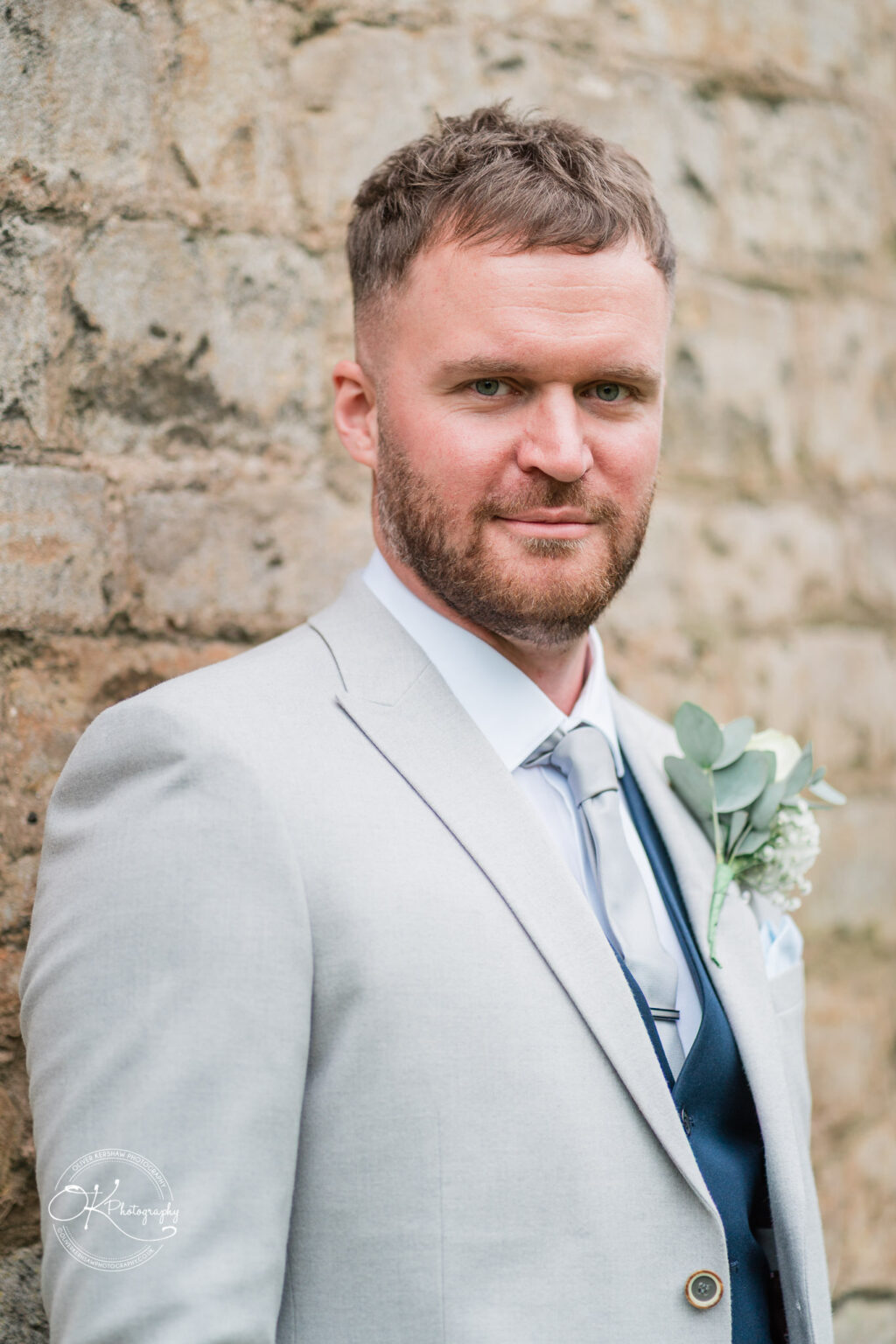 A man in a light grey suit and silver tie stands confidently against a stone wall, wearing a white flower boutonniere.