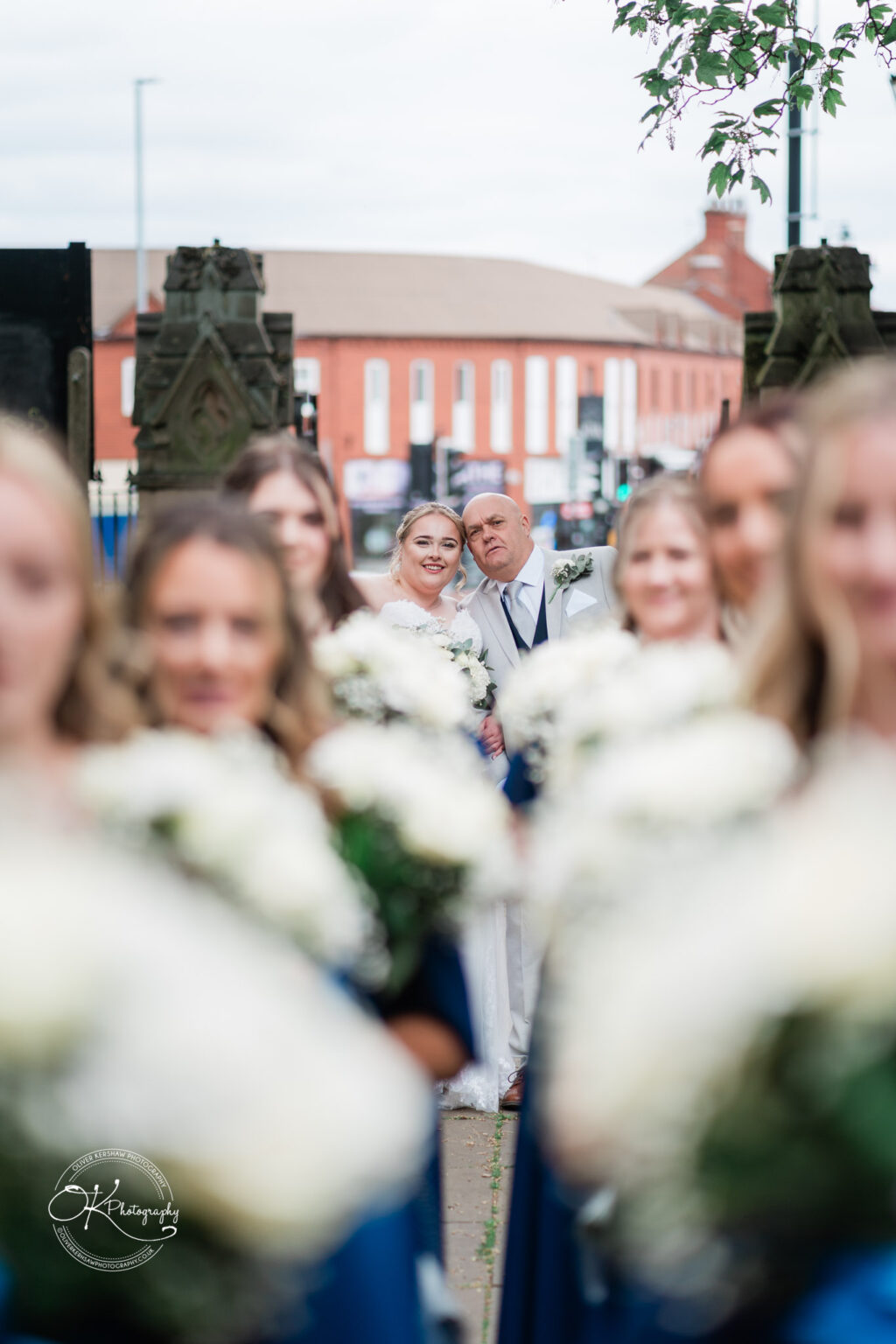 A bride and groom pose together, smiling in the background, framed by a group of bridesmaids holding white bouquets in the foreground.