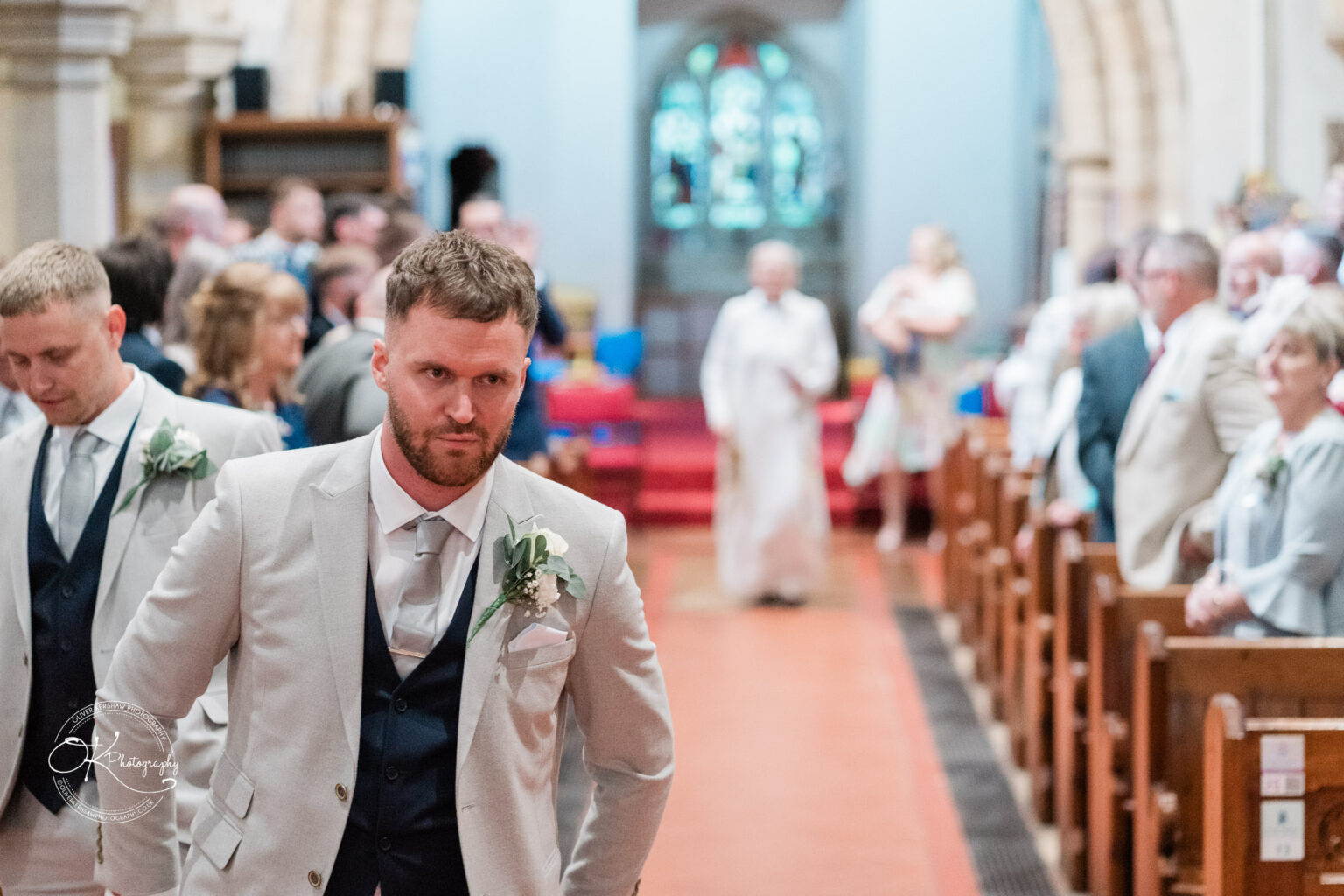 A focused groom in a light suit walks down the aisle of a church, with guests seated on either side, awaiting the ceremony.